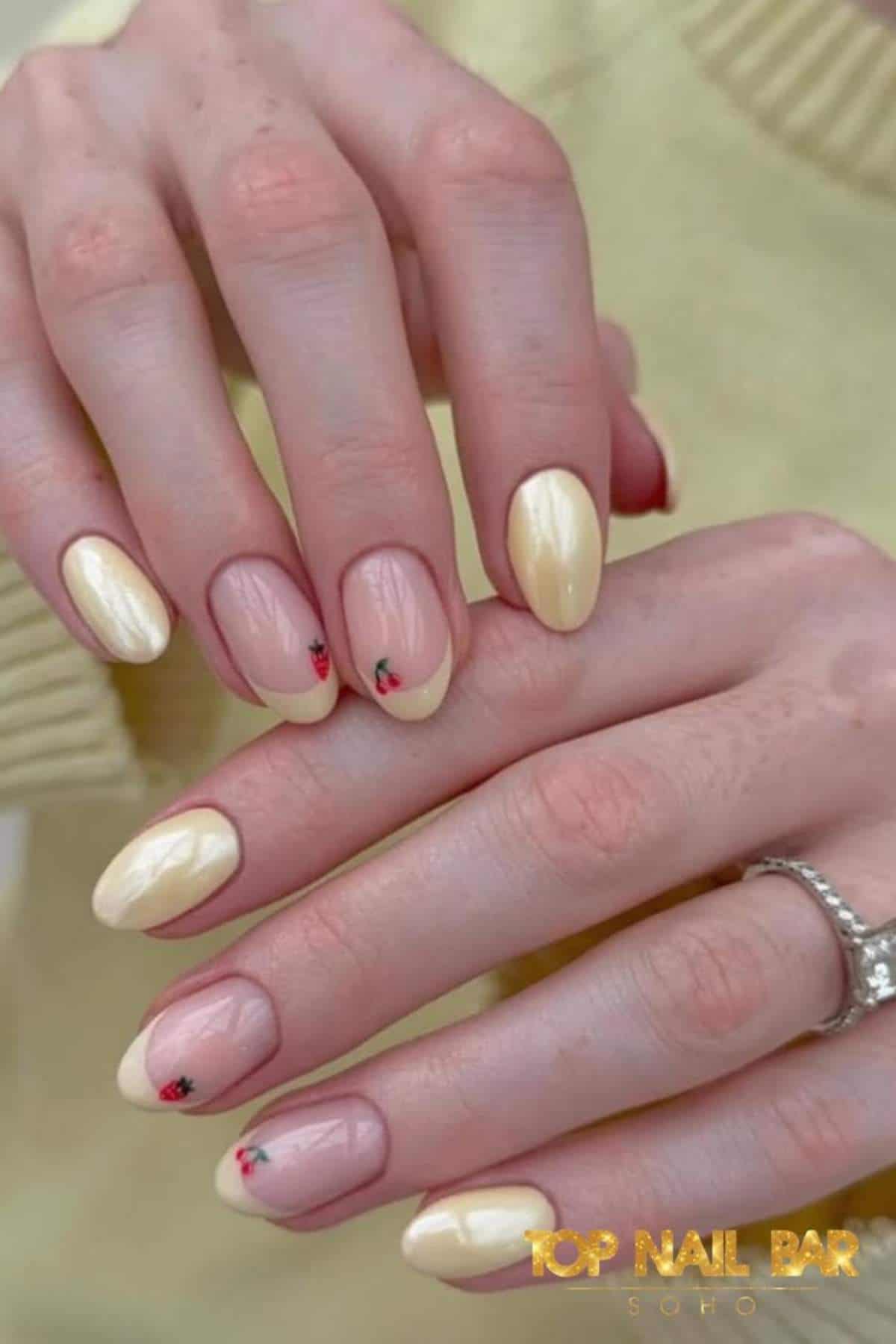 Hands with almond-shaped nails featuring a mix of pale yellow polish and nude polish with small red flower accents. A silver ring is worn on one finger.