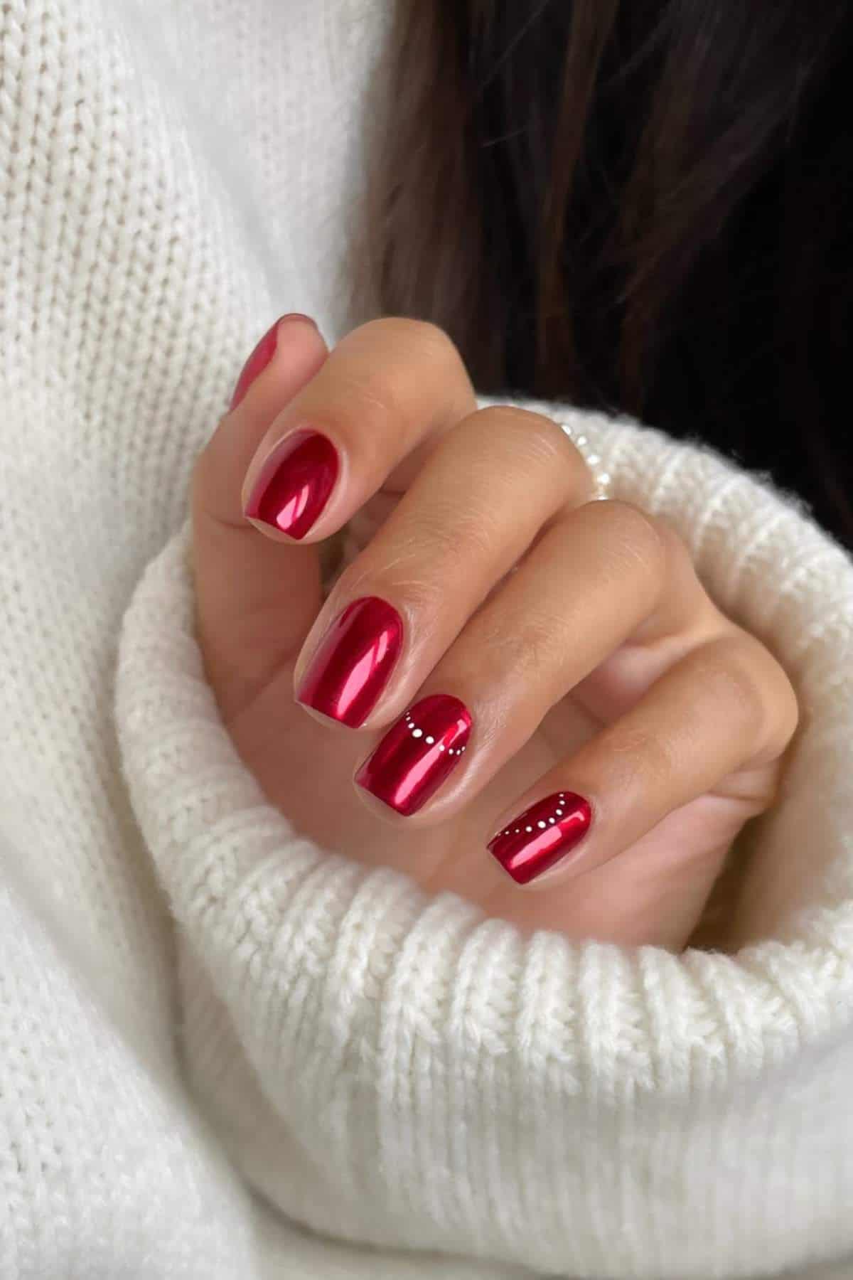A hand with shiny red nail polish and small silver studs on one nail, wearing a white knit sweater.
