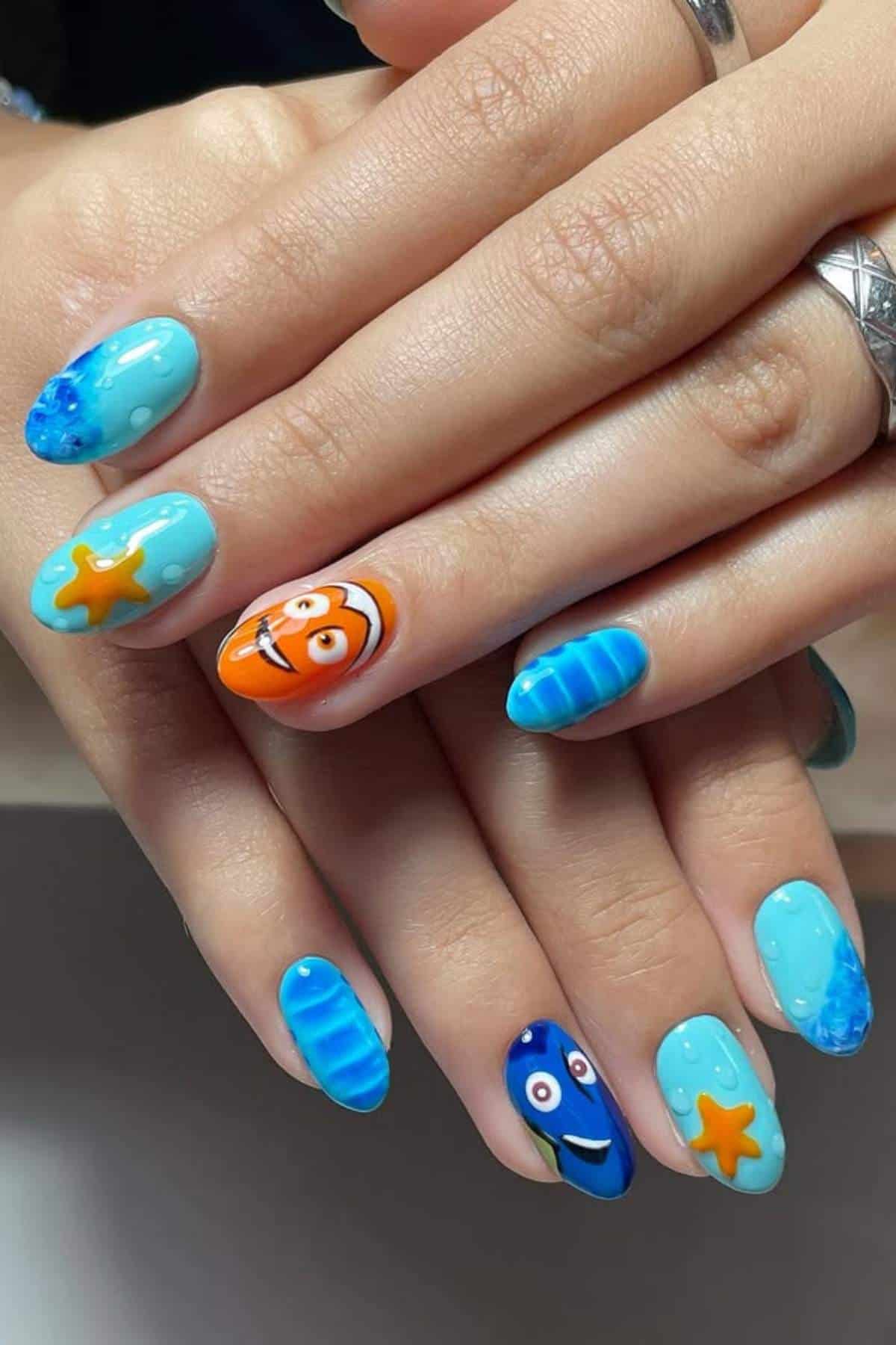 Hands with light blue manicure featuring cartoon fish, starfish designs, and ocean-like patterns on long, almond-shaped nails.