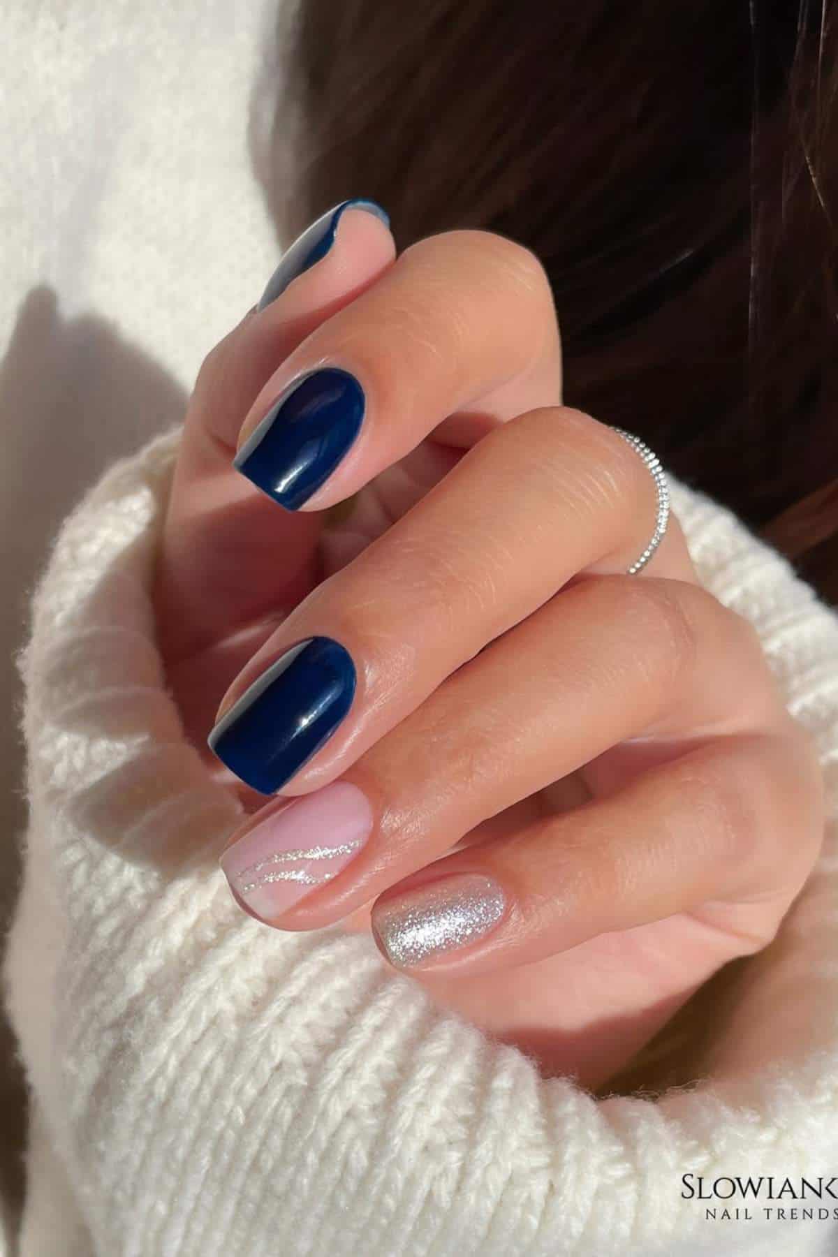 A hand with manicured nails featuring dark blue polish on three nails, one nail with silver glitter, and one with a clear base and a diagonal silver design.