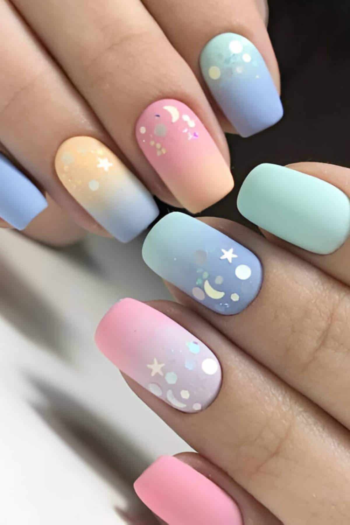 Close-up of hands with pastel gradient matte nails, decorated with small celestial designs including stars, moons, and dots.
