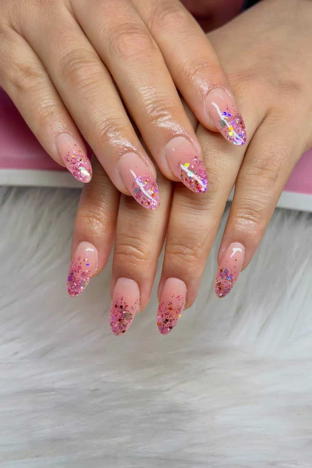 Hands with almond-shaped nails featuring pink glitter and star-shaped confetti nail art on a nude base, displayed on a light surface.