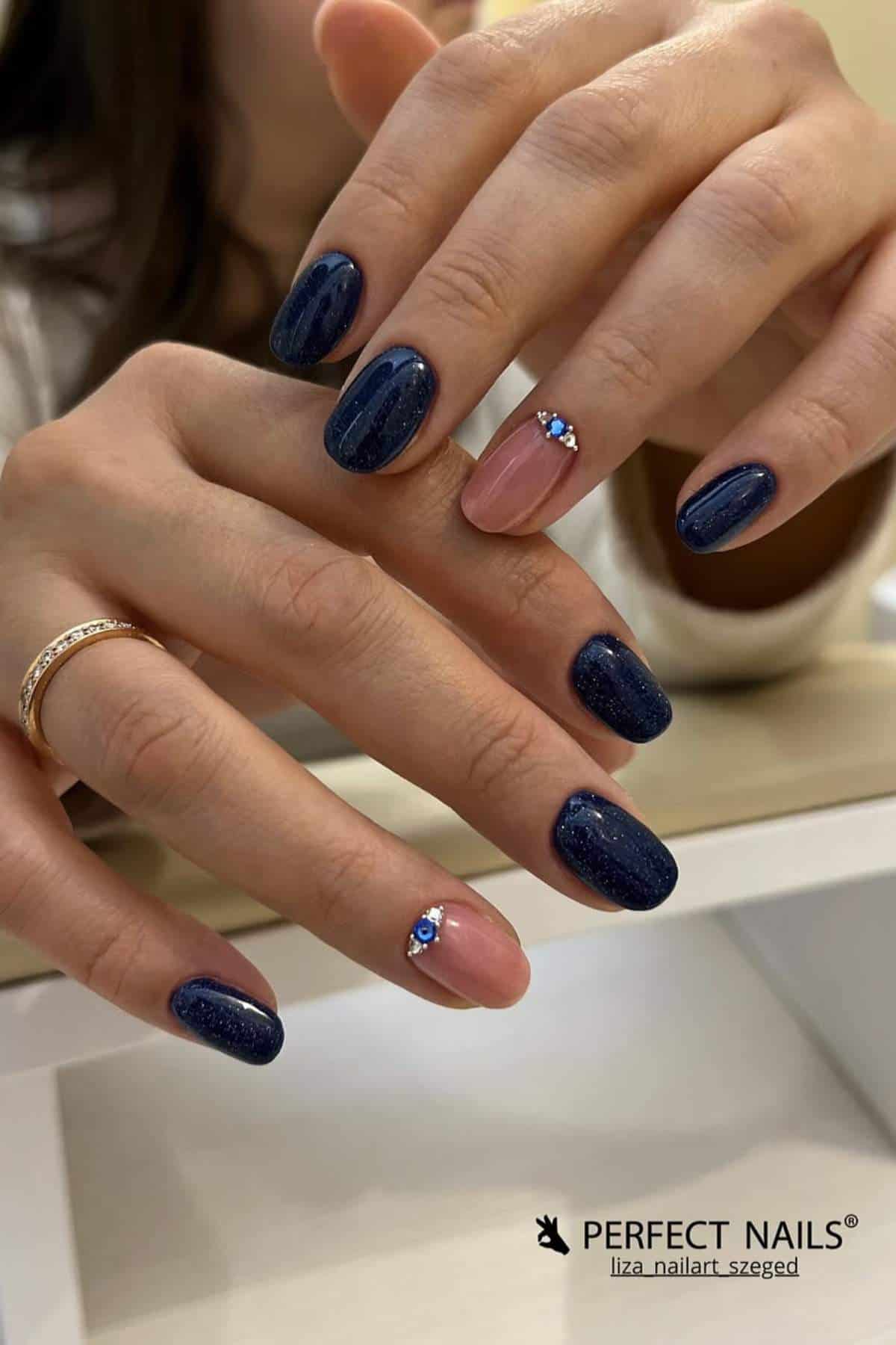 A woman displays a manicure featuring dark blue glittery nails and accent nails in light pink with small blue rhinestone decorations.