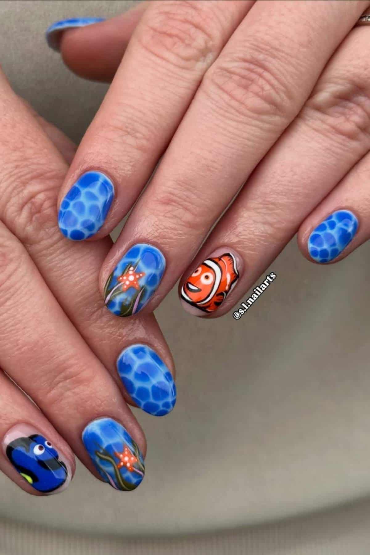 Close-up of hands with blue ocean-themed nail art featuring cartoon fish and seaweed, including designs inspired by Finding Nemo characters.