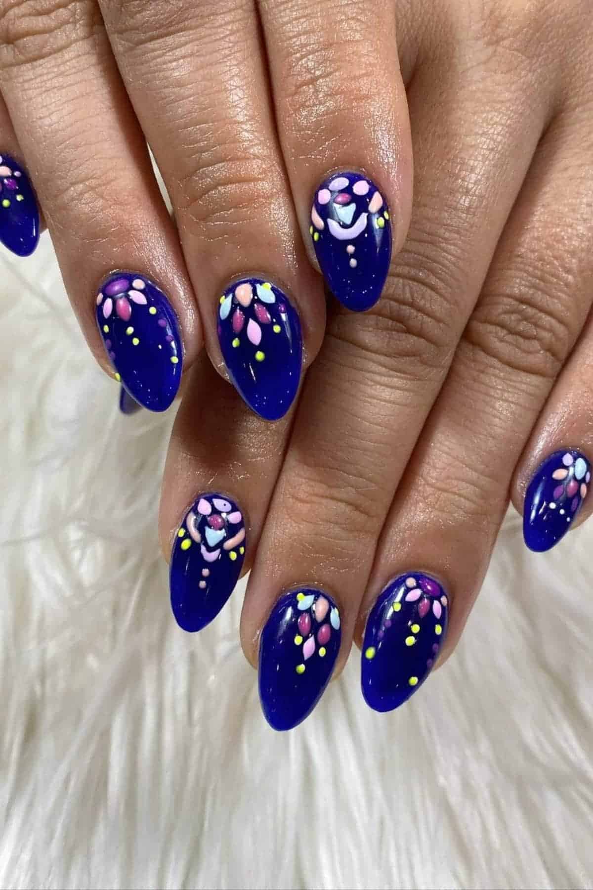 Hands with almond-shaped nails painted dark blue, featuring colorful floral and dot designs in pink, white, yellow, and light blue on a white textured surface.