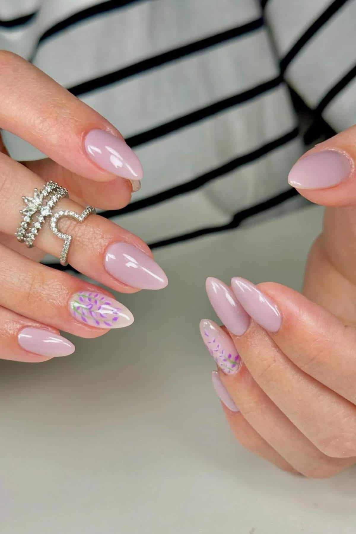 A person with almond-shaped, light pink manicured nails featuring small floral designs on two accent nails, wearing a silver ring, and a striped garment in the background.