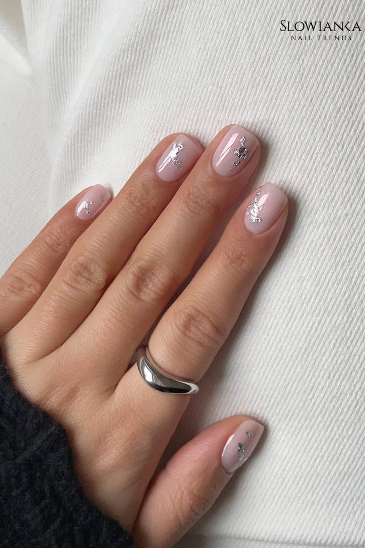 A hand with short, light pink polished nails featuring silver foil accents, resting on white fabric. The person wears a silver ring on the middle finger.
