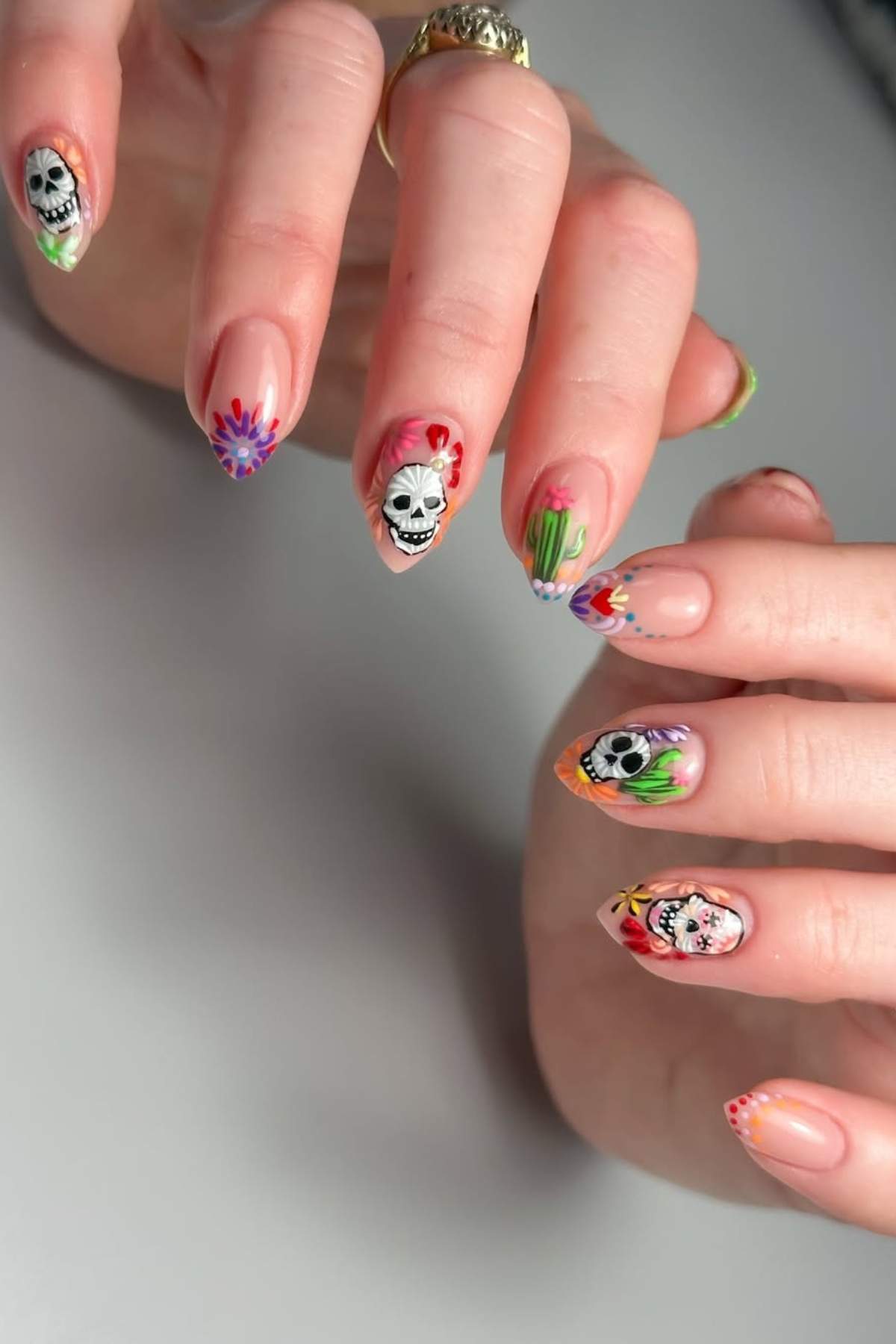 Hands with long, almond-shaped nails featuring colorful Day of the Dead skull designs and floral patterns on a light background.