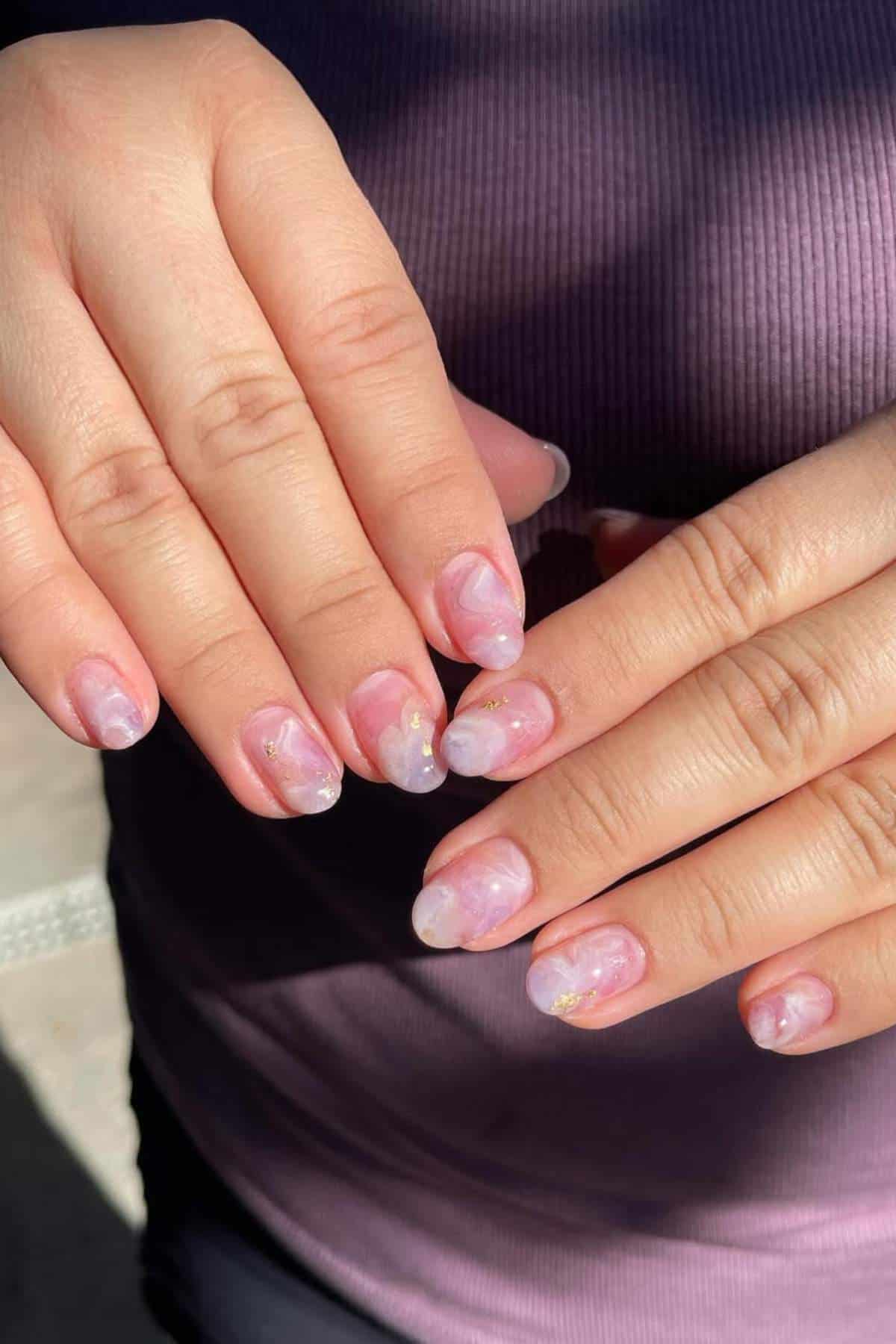 Hands with manicured nails featuring a soft pink and white marble design with subtle gold accents, against a ribbed purple shirt background.