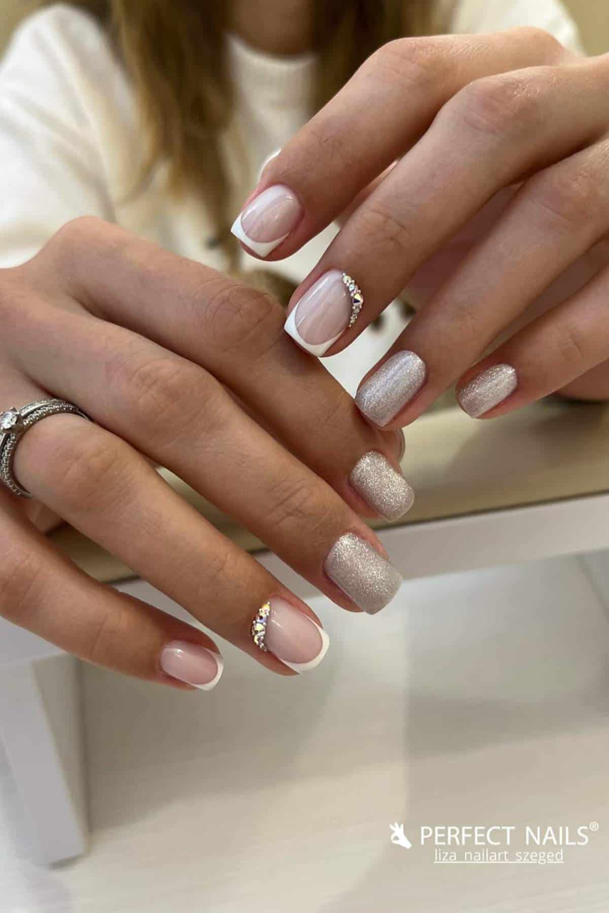 A person with manicured nails featuring a mix of French tips, glitter, and rhinestone accents rests their hands on a table.