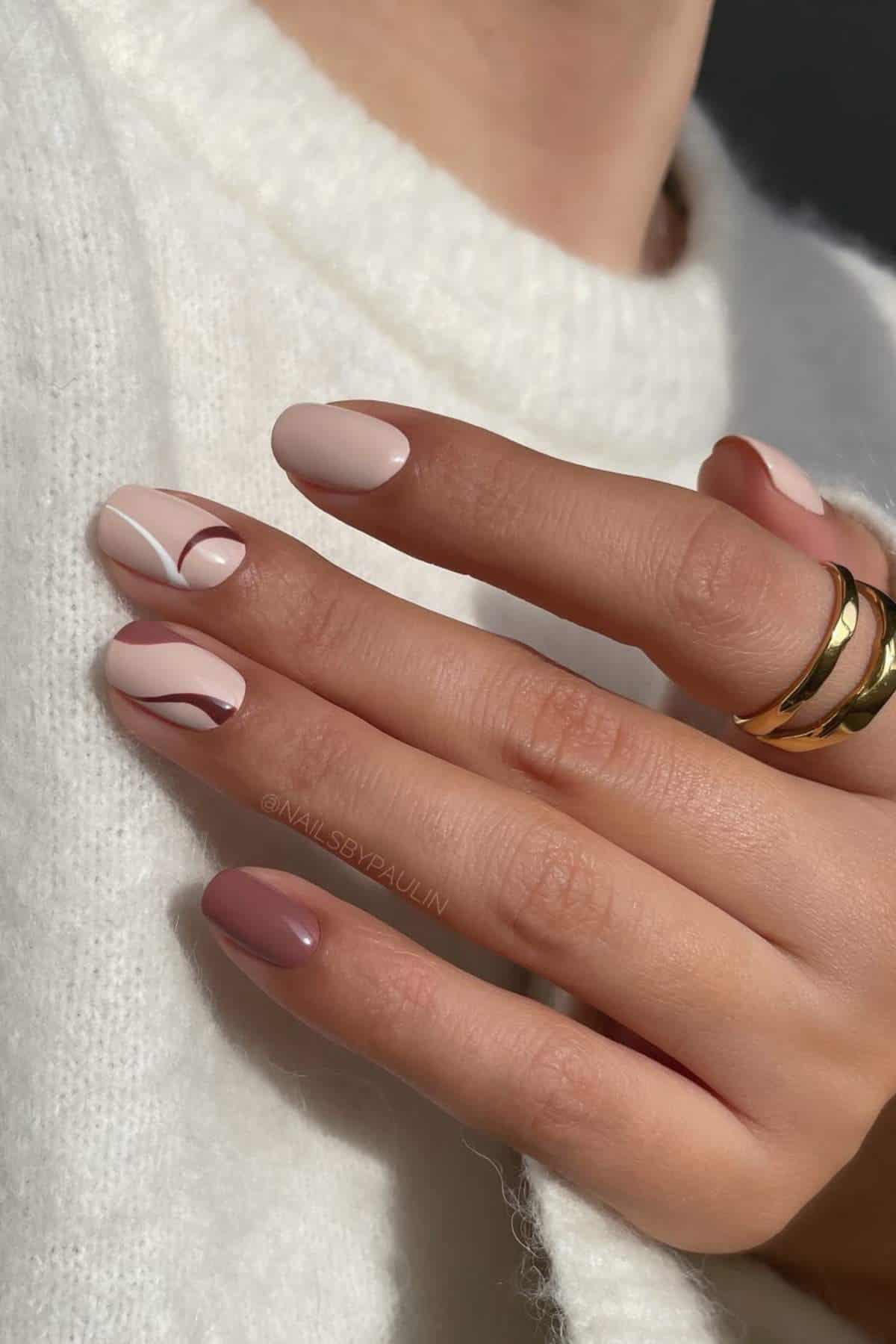 A hand with manicured nails in nude and brown tones featuring abstract designs, wearing two gold rings and a white knit sweater.