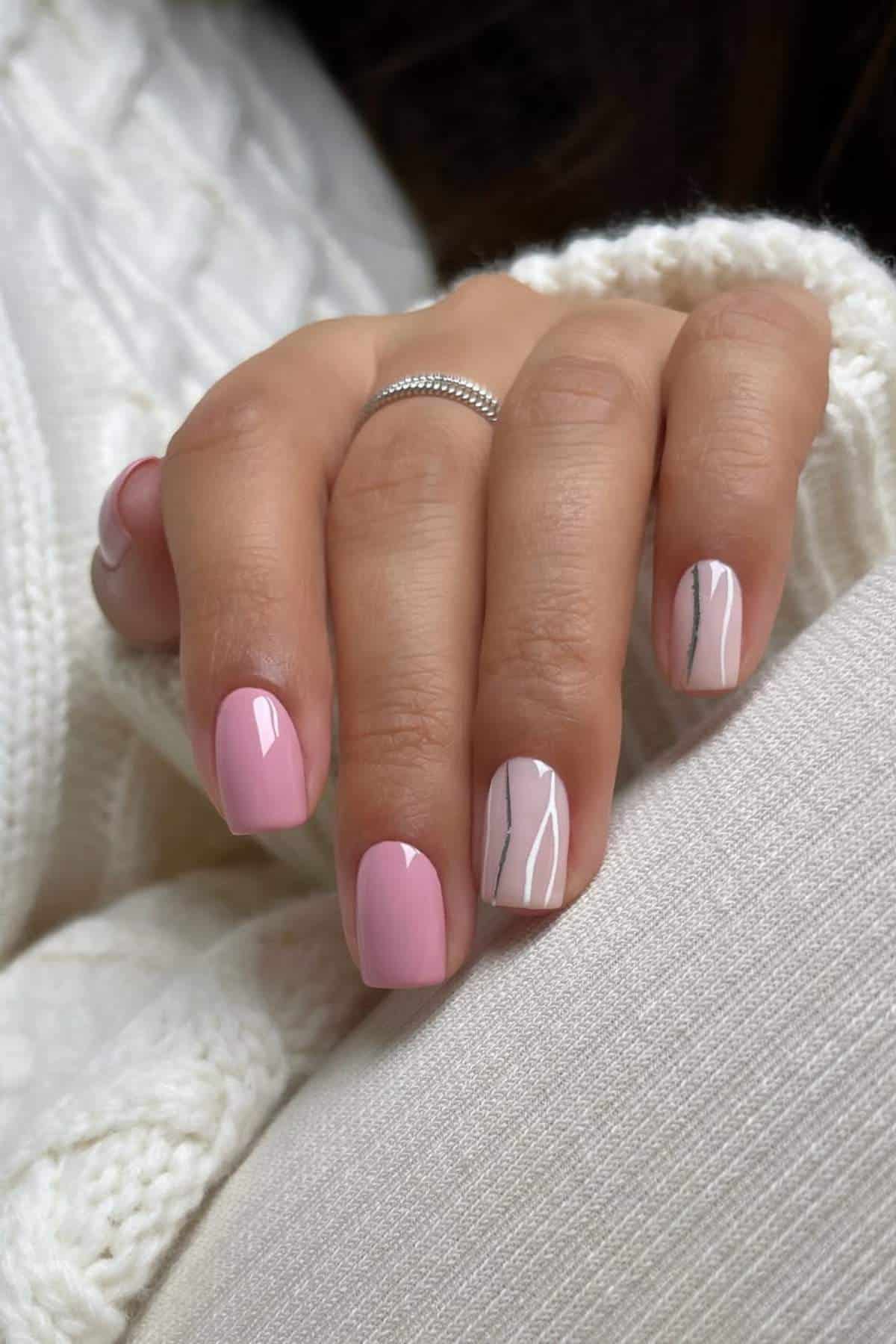 A hand with short, glossy light pink nails; two nails have simple silver line designs. The person wears a silver ring and a white knit sweater.