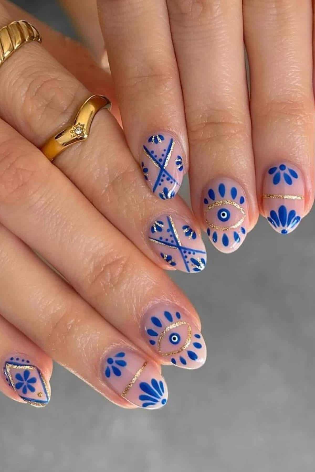 Close-up of hands with nude nails featuring intricate blue and gold geometric and floral nail art designs. The person is wearing gold rings.