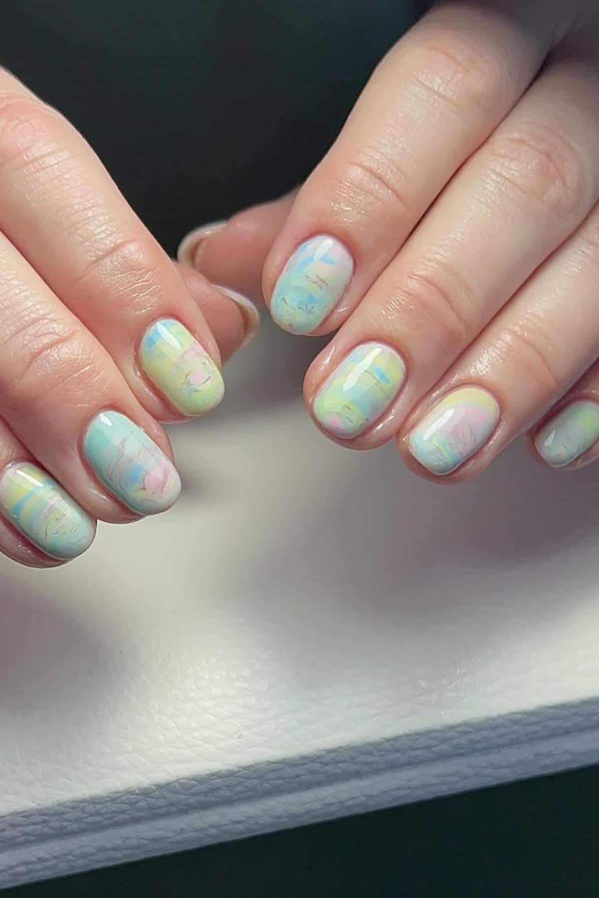 Hands with short nails painted in a pastel marble design featuring light blue, yellow, pink, and green shades.