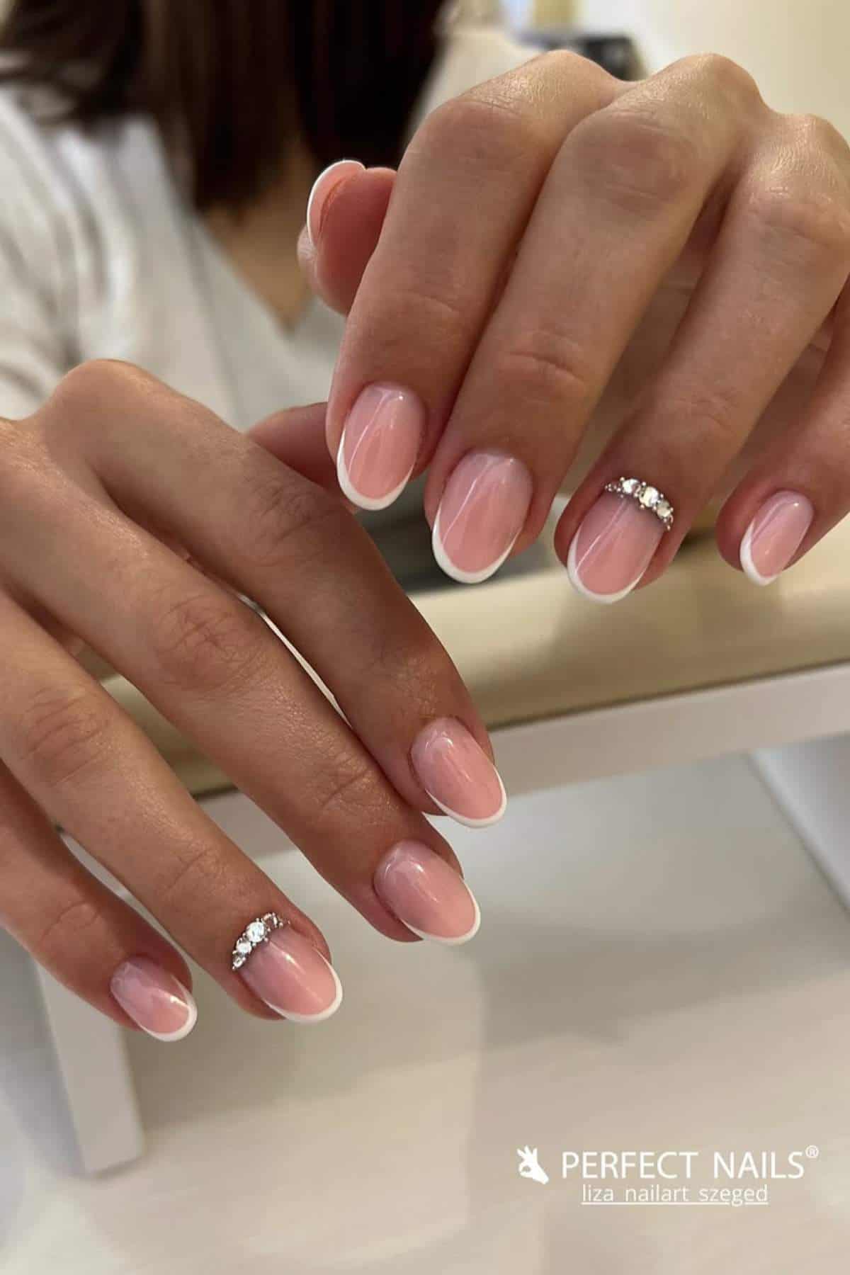 A person displays a French manicure with a pale pink base, white tips, and rhinestone accents on two ring fingers.