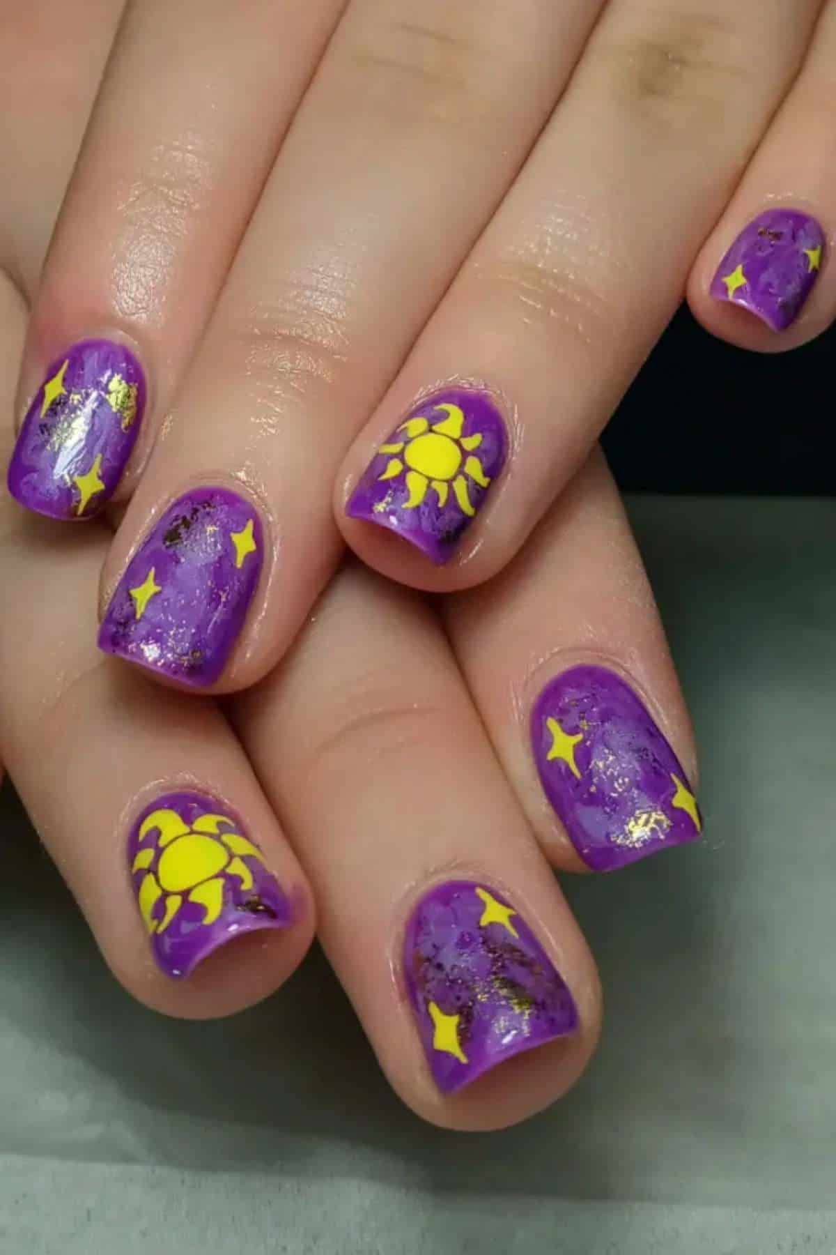 Hands with short nails painted purple and decorated with bright yellow sun and star designs.
