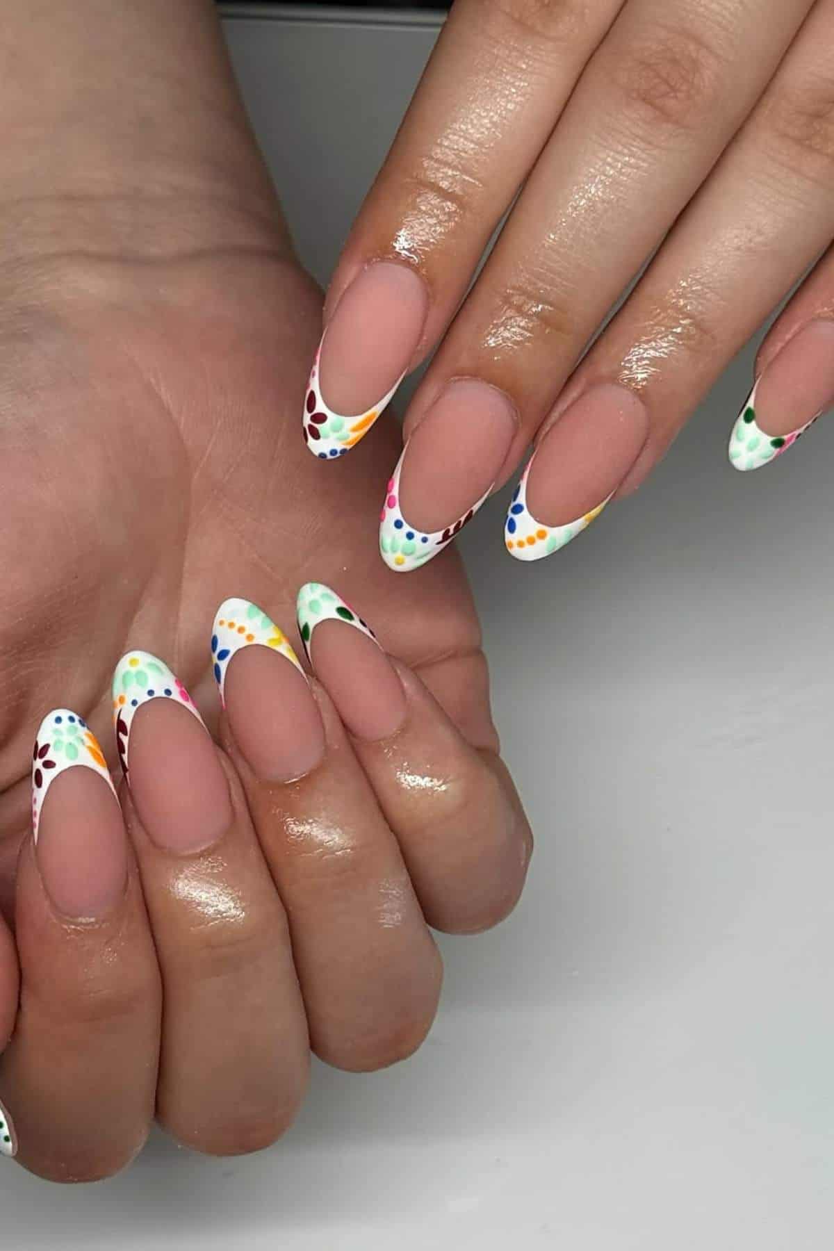 Hands with almond-shaped nails featuring a nude base and French tips decorated with colorful polka dot patterns in various shades.