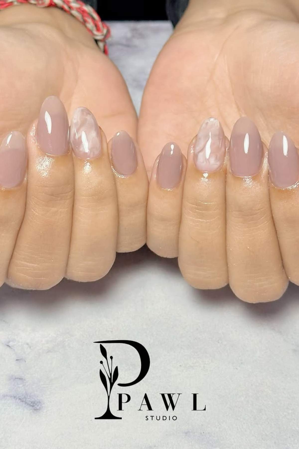 Hands with almond-shaped nails painted in glossy nude tones, with a marble-like white accent design on the ring fingers. "PAWL Studio" logo visible at the bottom.