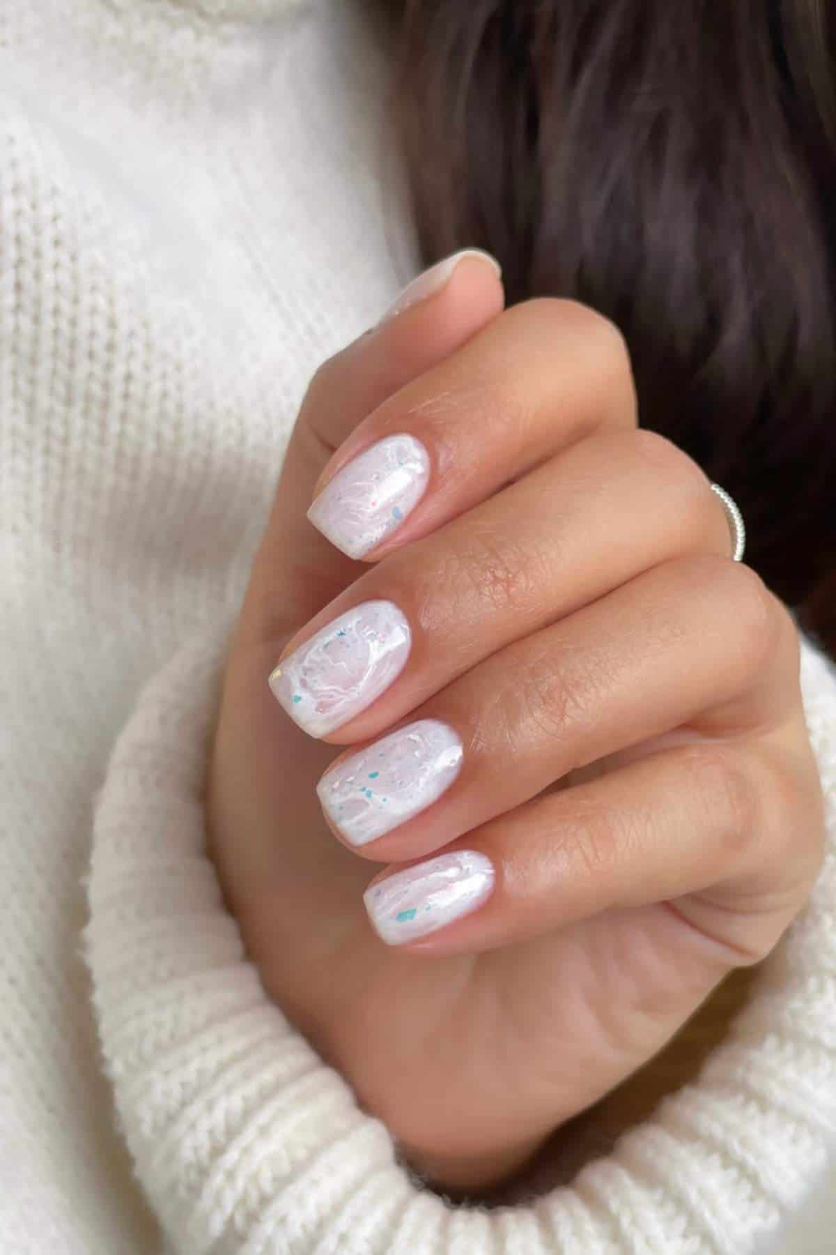 A hand with square-shaped fingernails painted in a glossy, pale pink polish with subtle white marbling and shimmer, against a cream knit sweater.