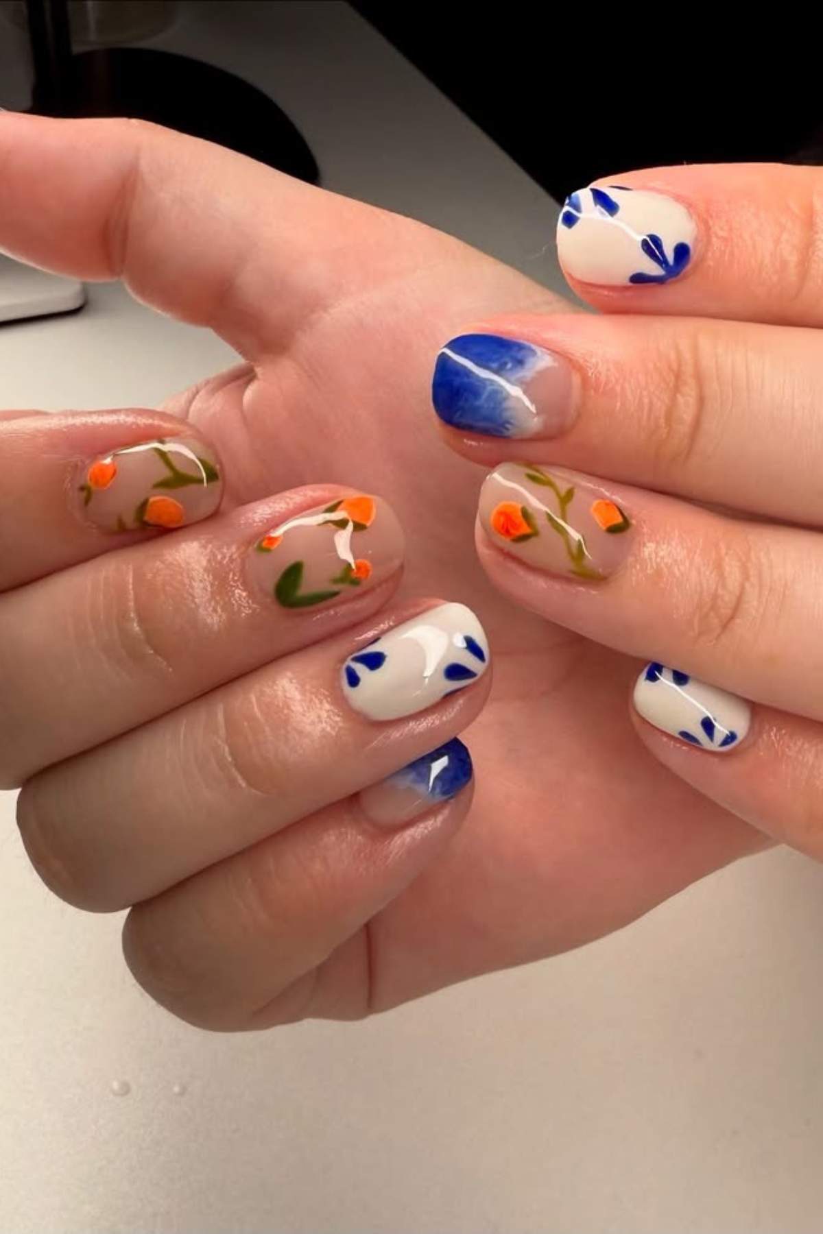 A hand with manicured nails featuring blue abstract designs, orange and green floral patterns, and neutral-colored polish on a light background.