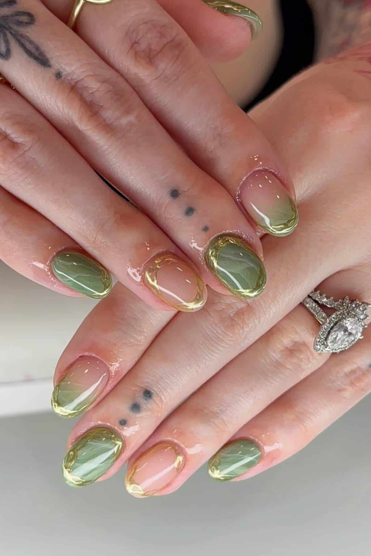 Close-up of hands with green and nude gradient manicure, gold outlines on the nails, tattoos on fingers, and a diamond ring on one finger.