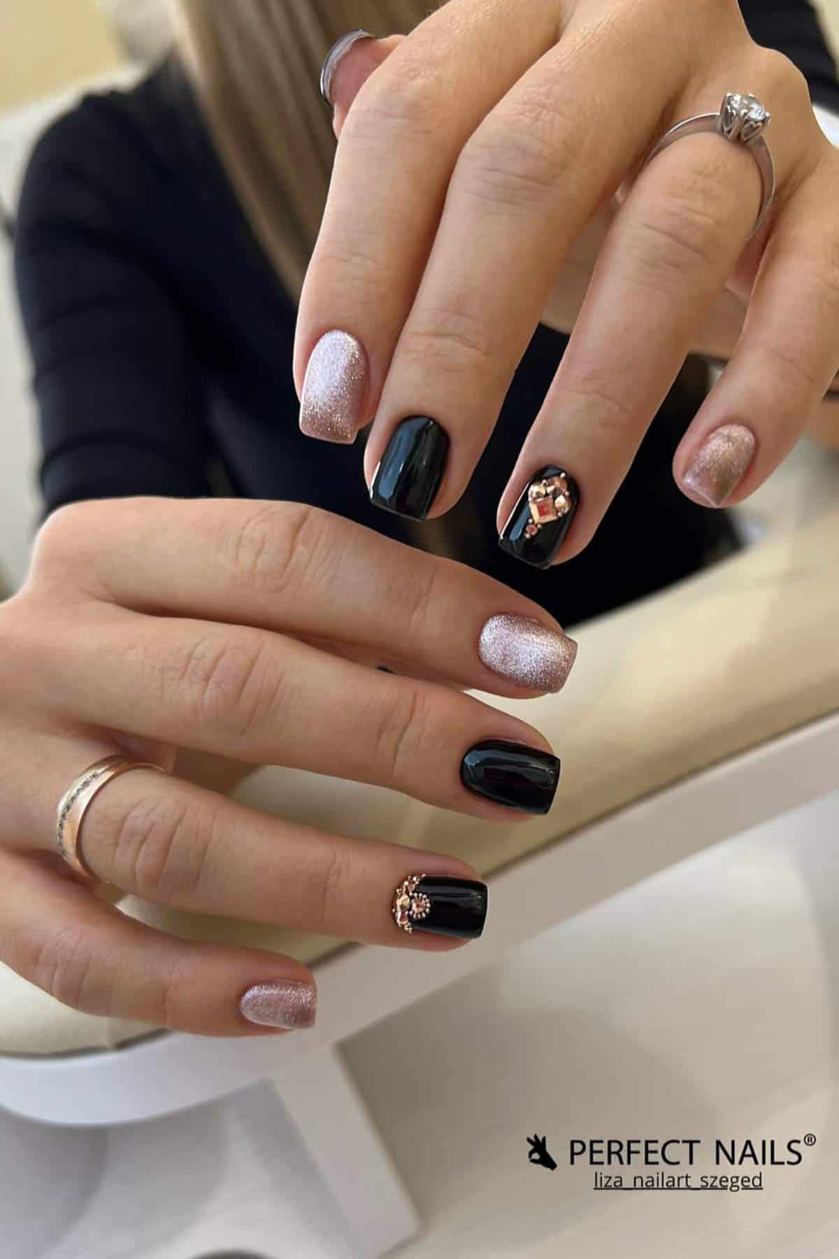 Close-up of hands with manicured nails featuring black polish, glitter accents, and decorative gems; person is wearing rings on several fingers.