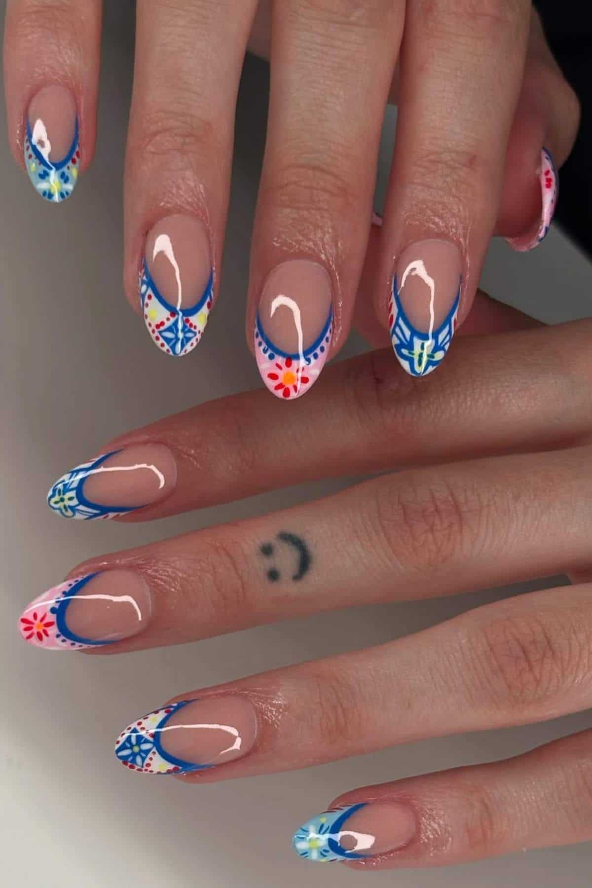 Close-up of hands with long, almond-shaped nails featuring colorful, intricate geometric nail art and a small smiley face tattoo on one finger.