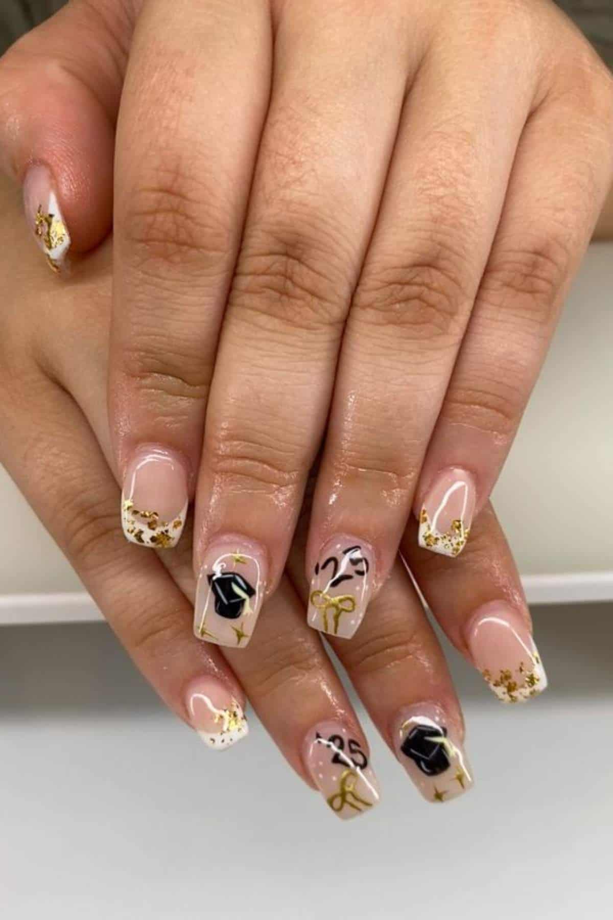 Hands with manicured nails featuring gold glitter tips, black graduation cap designs, the number "23," and gold diploma scrolls on a nude base.