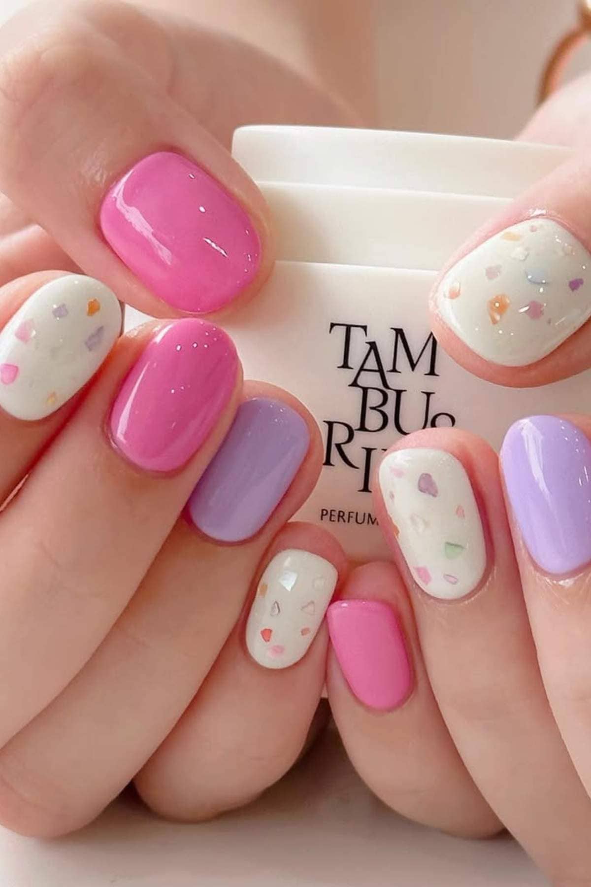 Close-up of hands with manicured nails painted in pink, purple, and white with colorful confetti accents, holding a white container with black text.