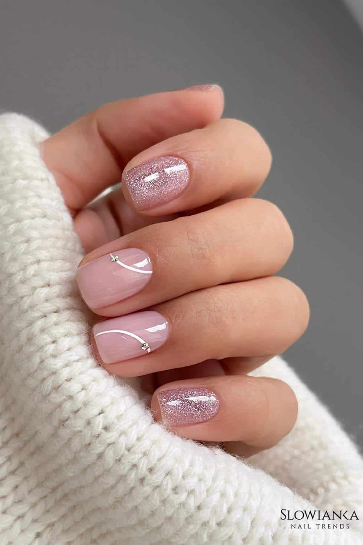 A hand with short, almond-shaped nails featuring light pink polish, silver glitter accents, and minimalistic rhinestone decorations, resting on a cream knitted fabric.