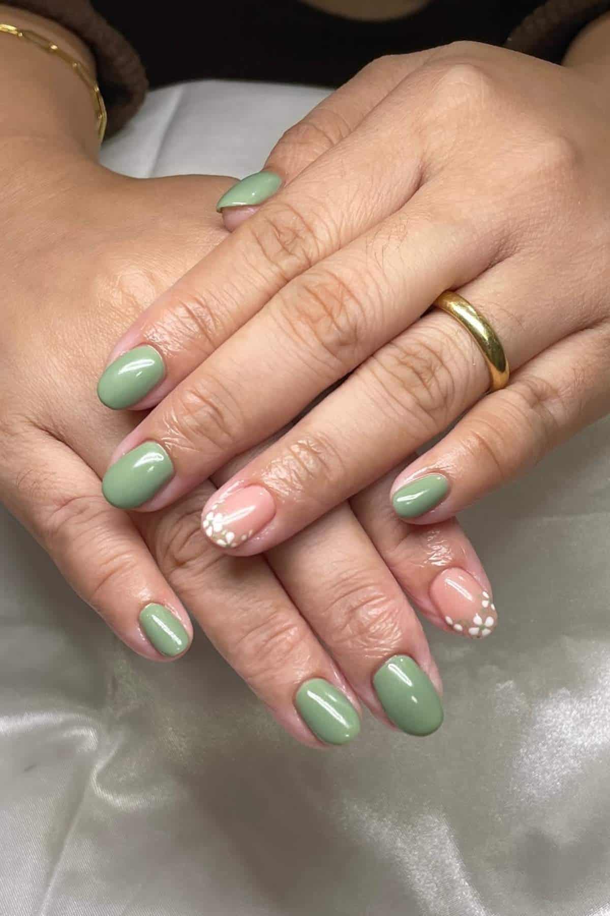 Hands with oval-shaped nails, most painted green; ring fingers feature a nude base with white floral designs. Gold ring on one finger.