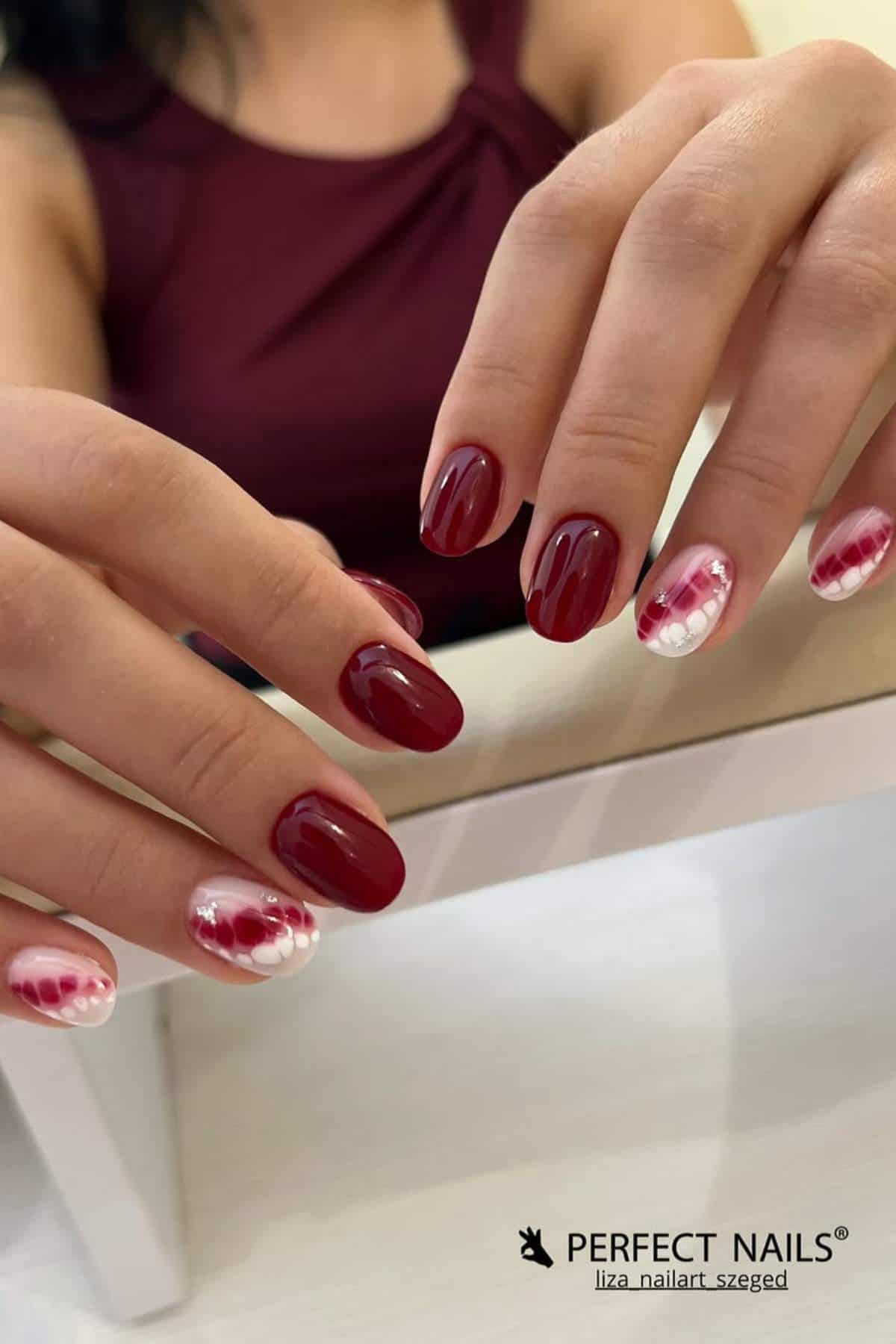 A person displays a manicure with deep red polish and white-red gradient designs with dots on two accent nails. The person wears a sleeveless burgundy top.