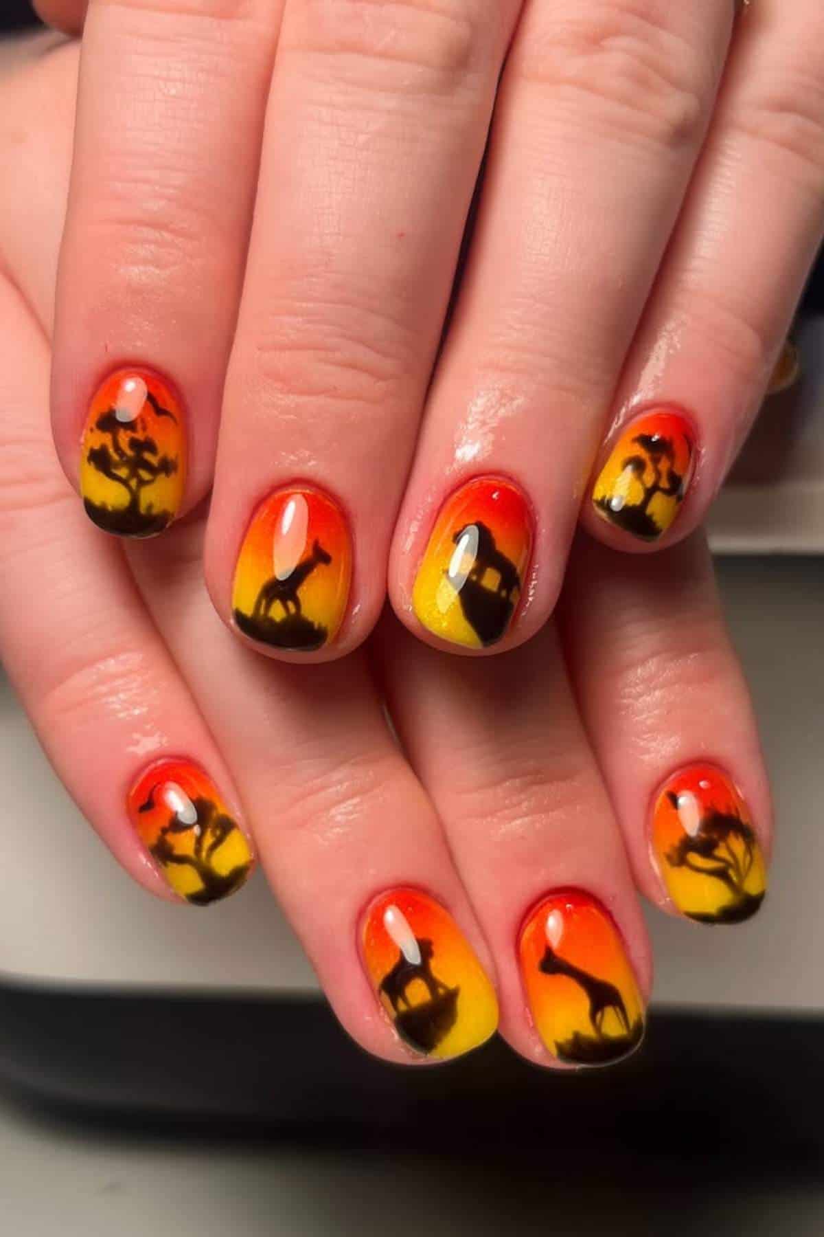 Fingernails painted with an orange and yellow gradient, featuring black silhouettes of African wildlife and trees, resembling a sunset savanna scene.