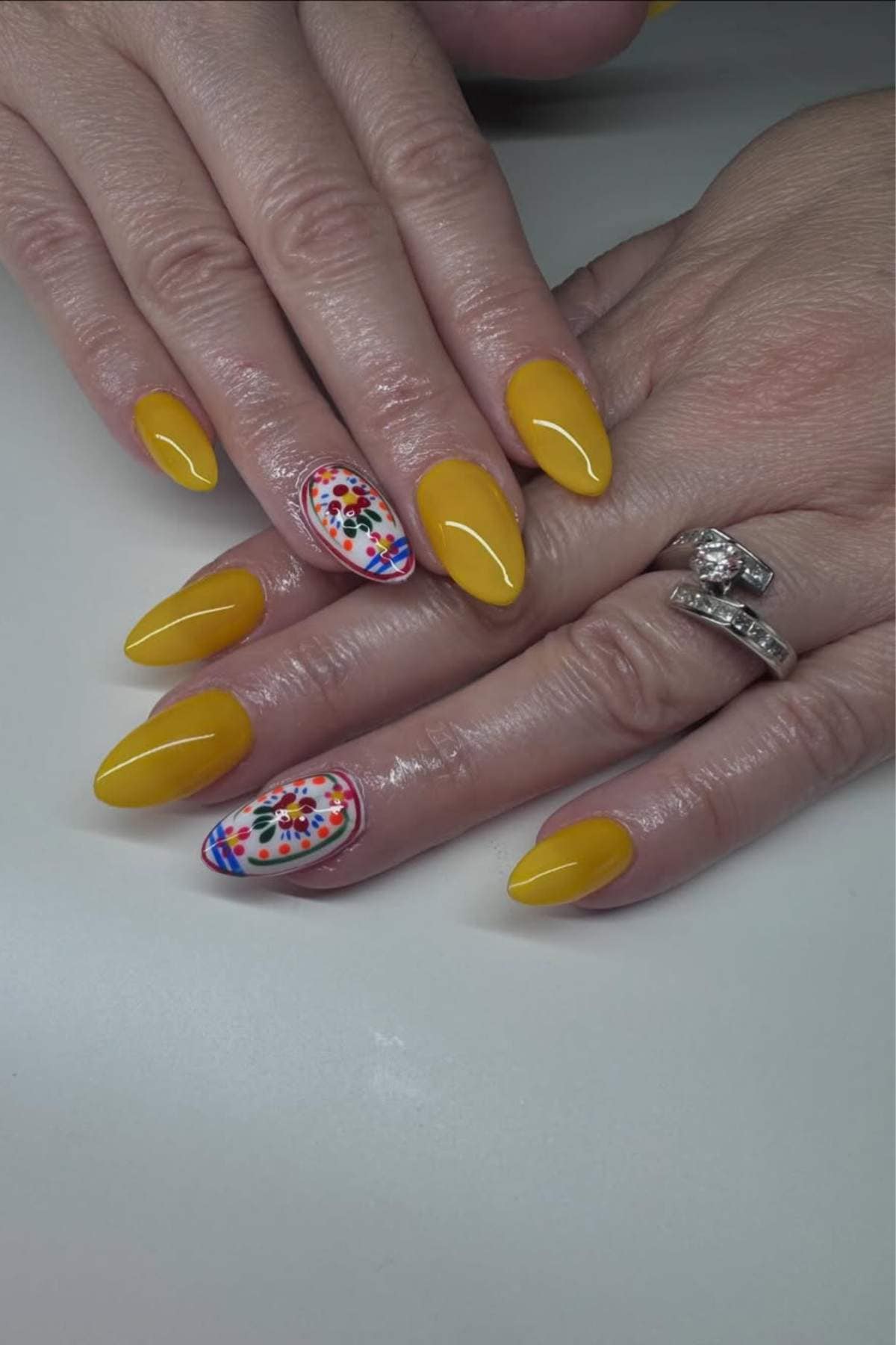 A hand with yellow almond-shaped nails, except one accent nail on each hand featuring a colorful floral design.