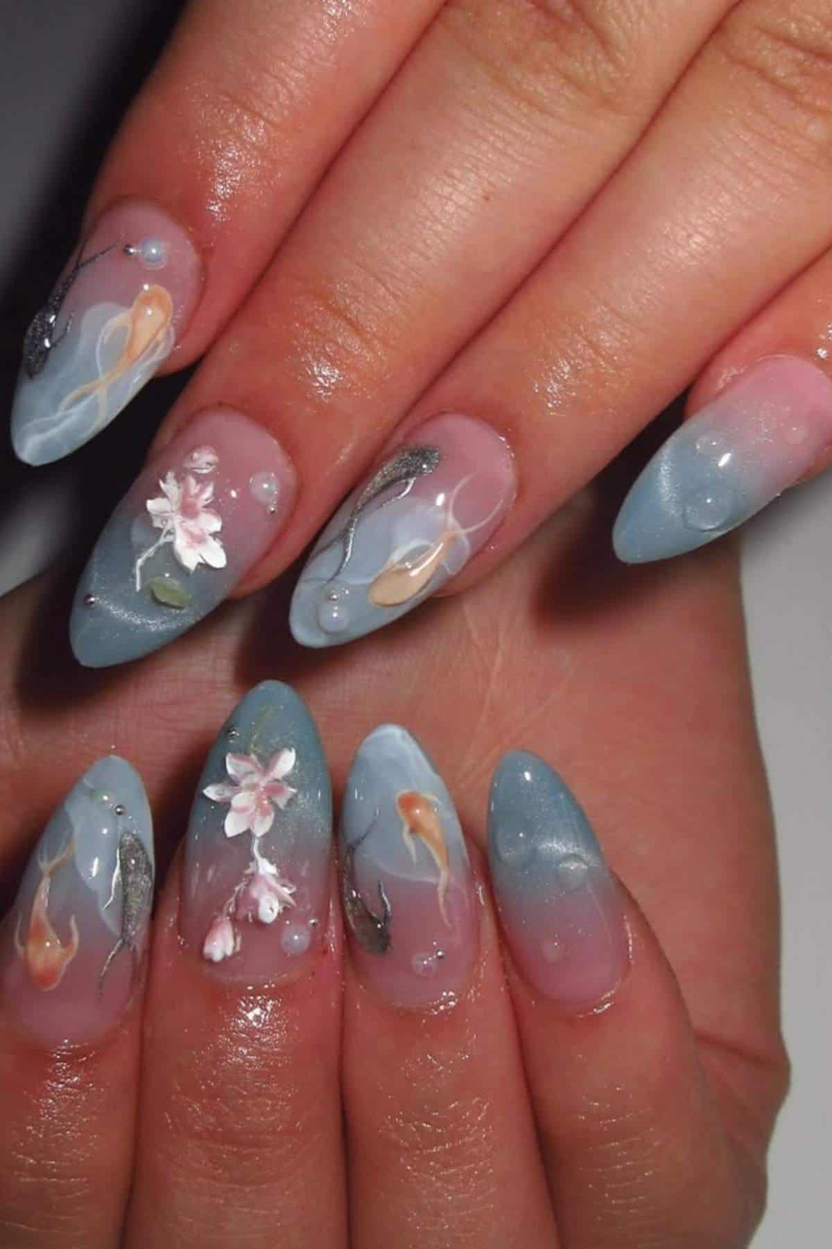 Hands with long, almond-shaped nails featuring blue and pink ombré polish, delicate 3D white floral designs, and painted fish-like accents.