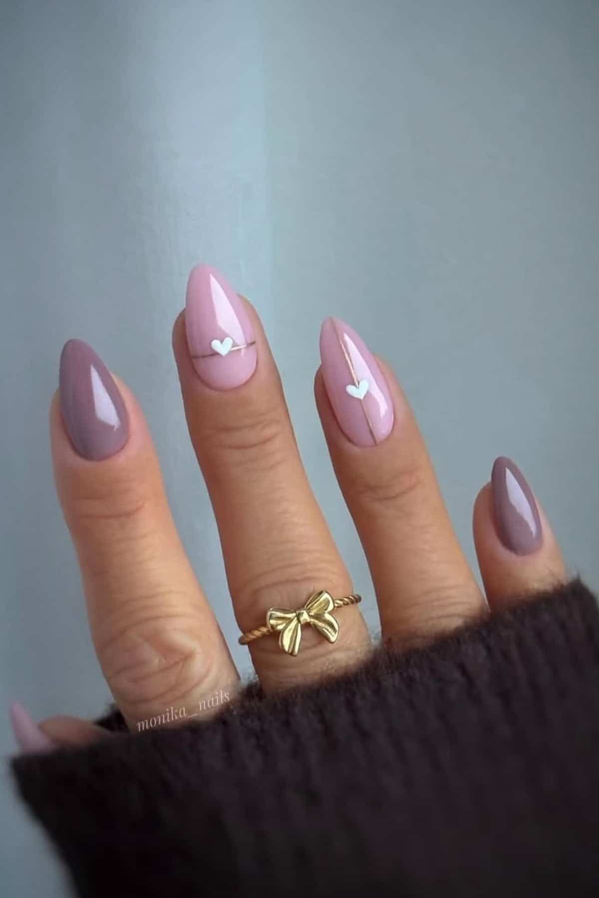 A hand with almond-shaped nails painted in mauve and light pink, featuring small white heart decals on two nails, wearing a gold bow ring.