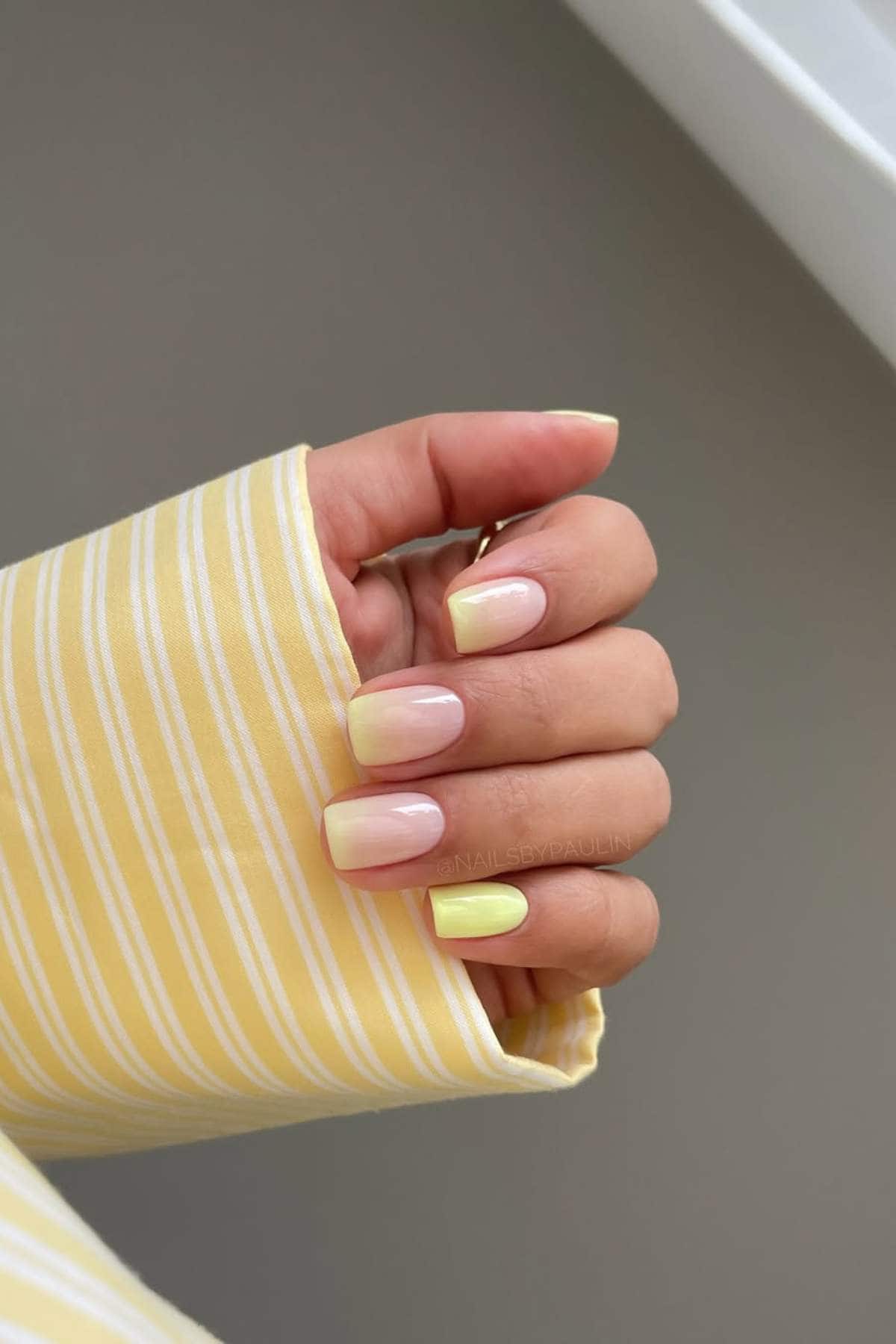 A hand with square-shaped nails painted in a pastel yellow and nude ombre design, shown against a yellow and white striped sleeve.