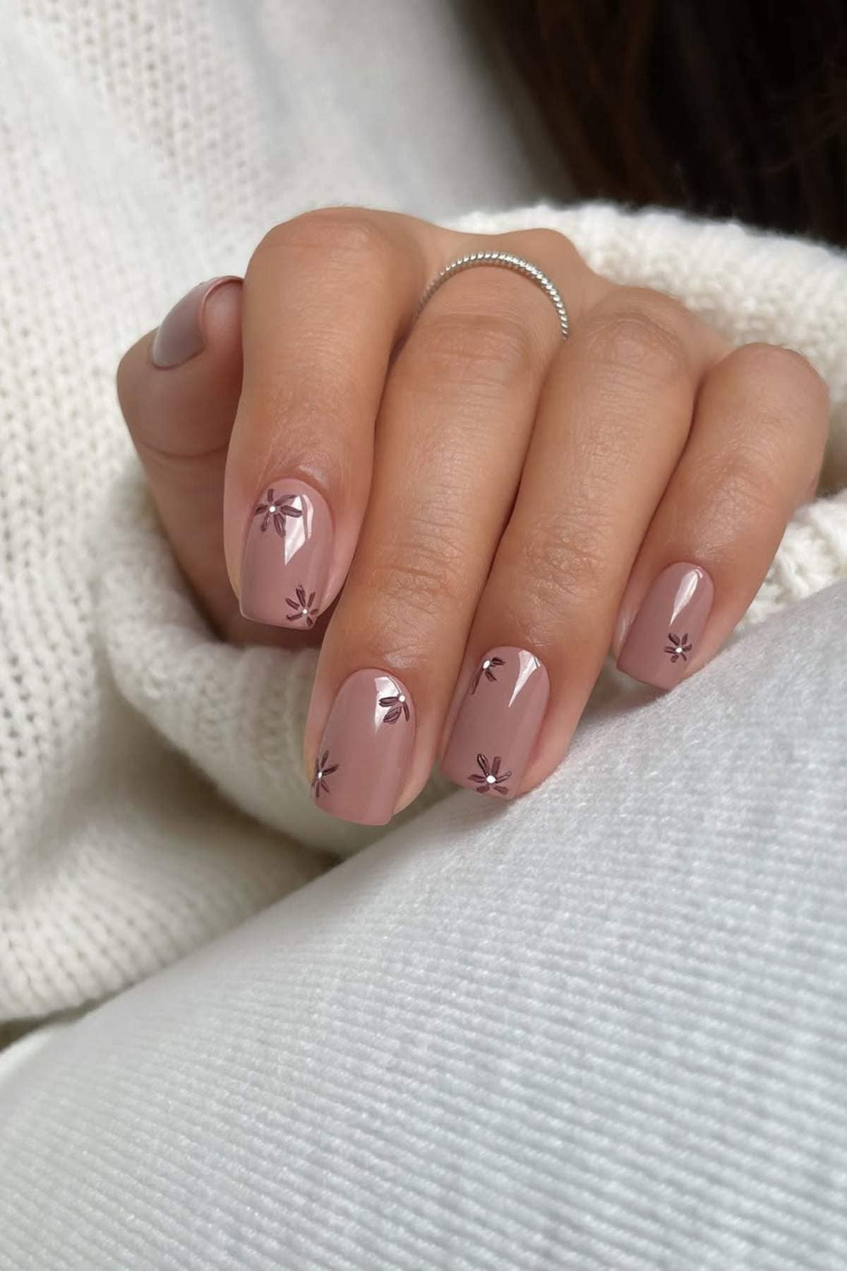 A hand with short, nude-colored nails featuring small, dark floral designs. The person is wearing a silver ring and a white knit sweater.