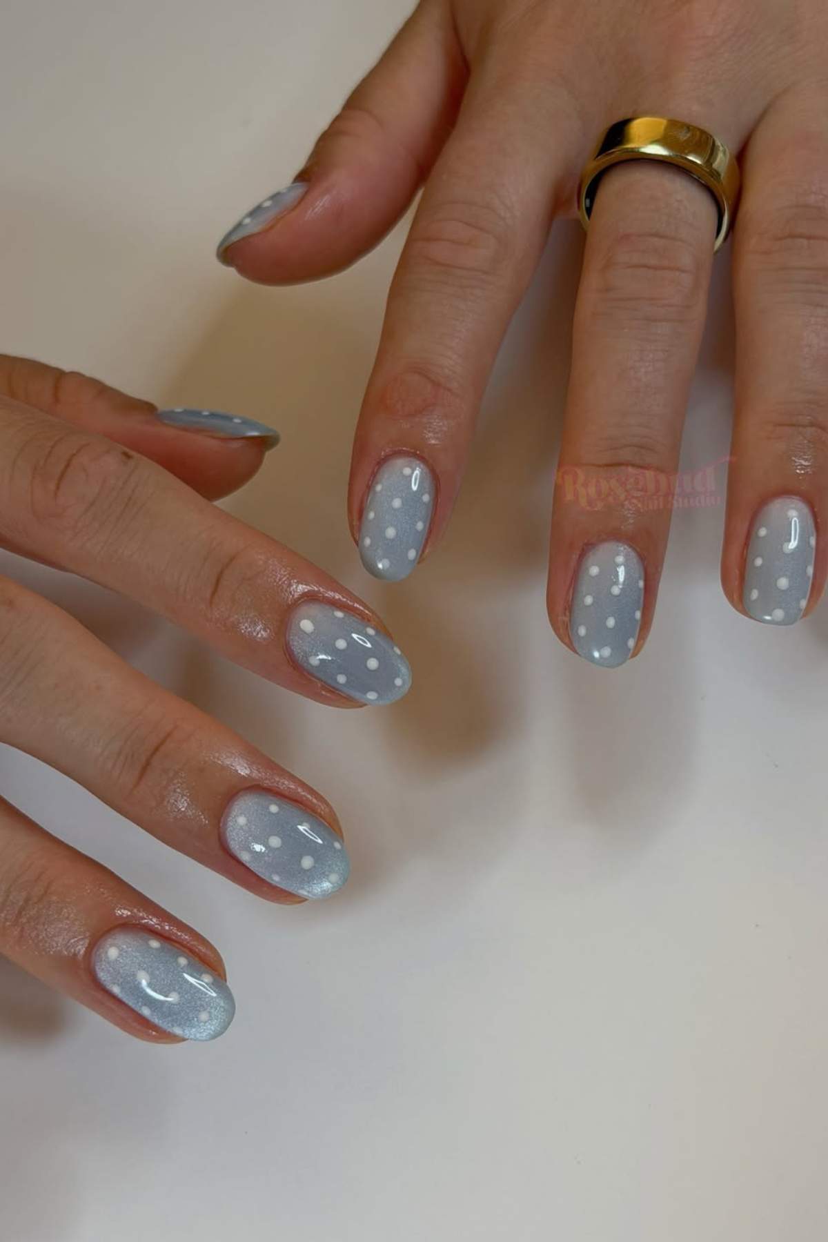 Hands with light blue, glossy nail polish featuring white polka dots. One hand has a gold ring on the ring finger.