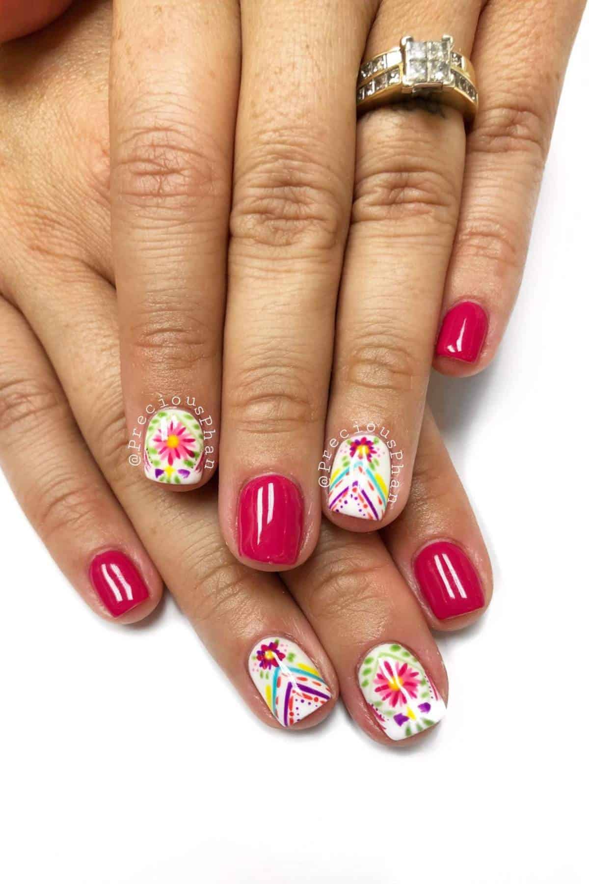 Hands with bright pink nail polish; ring fingers feature colorful floral and geometric nail art. The person is wearing two gold rings with a prominent diamond.