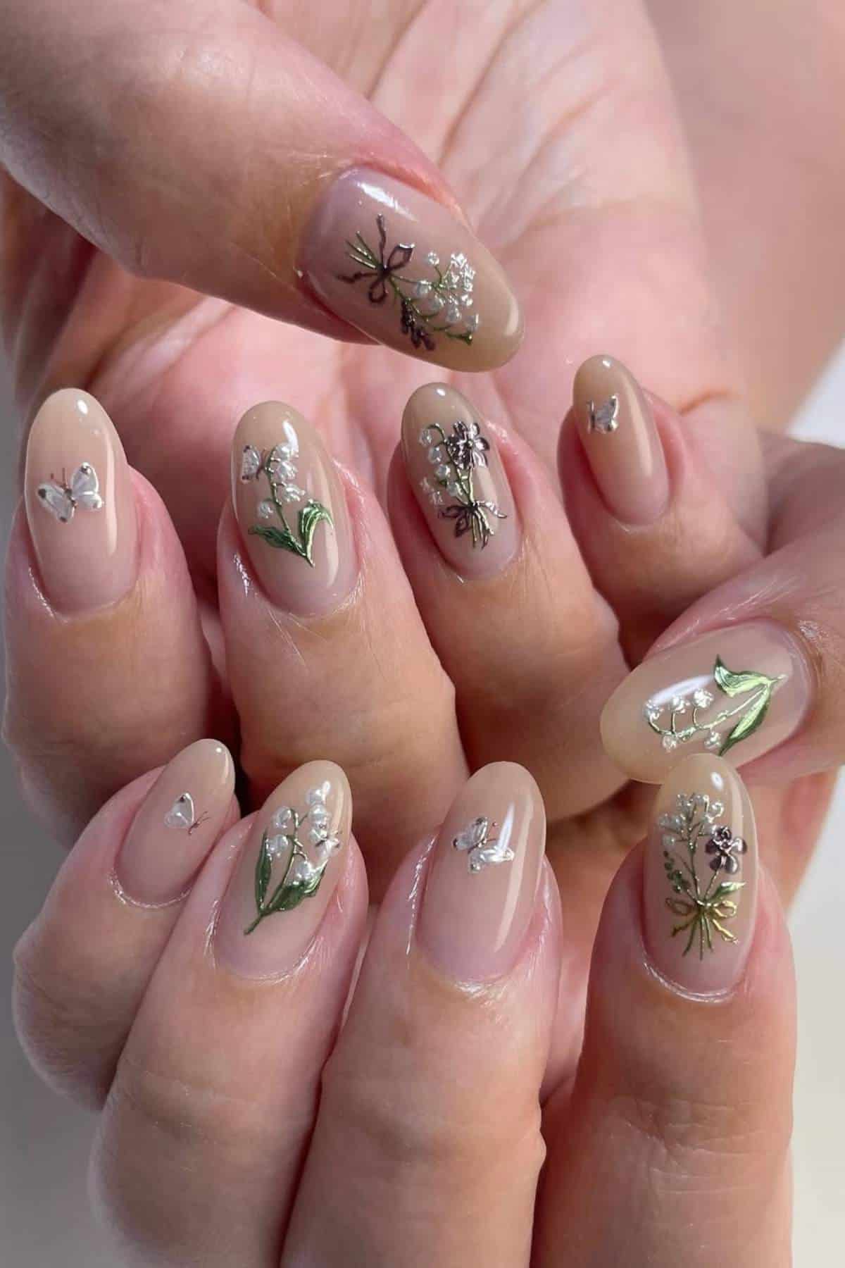 Close-up of hands with almond-shaped nails painted nude and decorated with small white flowers, green leaves, and silver butterfly designs.
