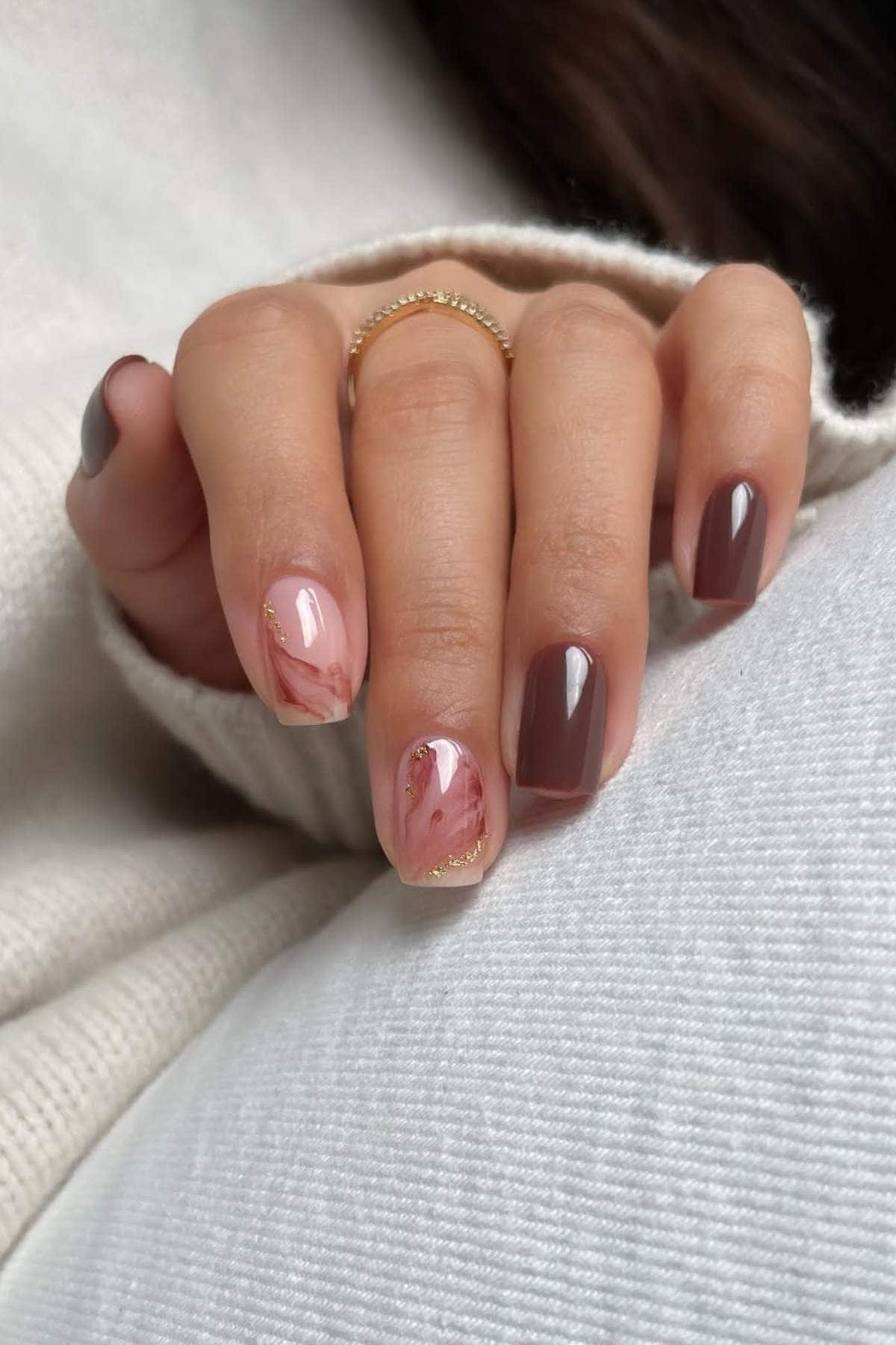 Hand with manicured nails featuring brown polish, pink marble accent nails, and gold detailing, resting on a light-colored fabric. A gold ring is worn on one finger.