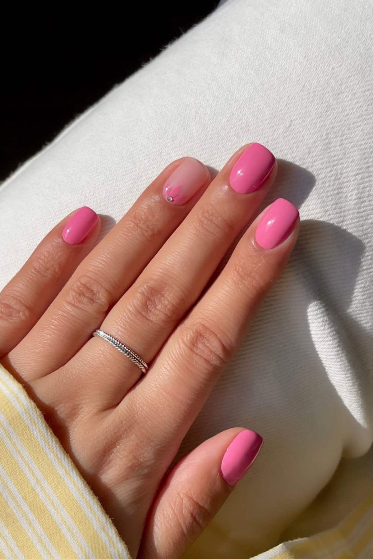 A hand with short, rounded nails painted pink, except for one natural nail with a small gem accent. The person wears a thin silver ring and rests their hand on white pants.