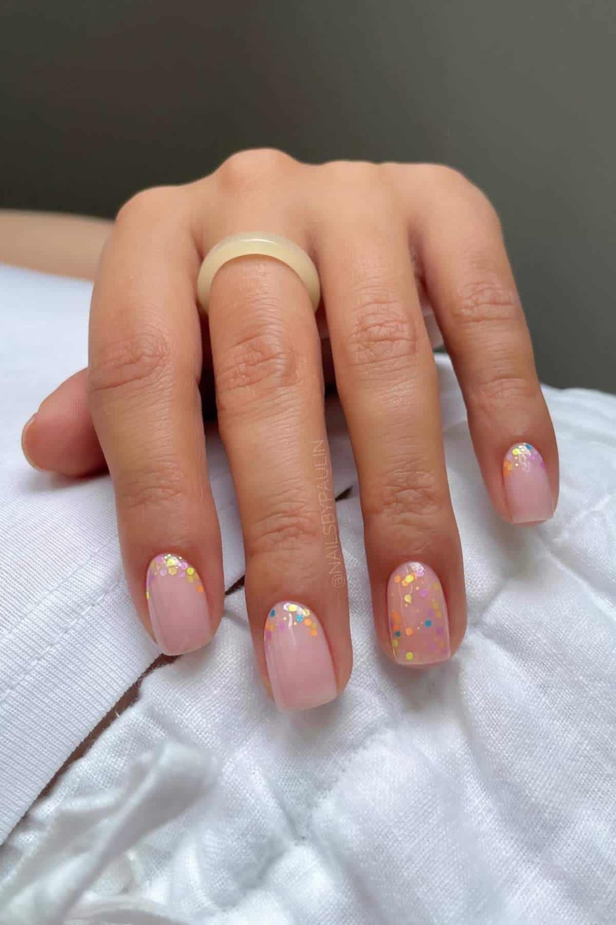 A hand with short, rounded nails painted light pink and decorated with colorful glitter, resting on a white fabric surface.