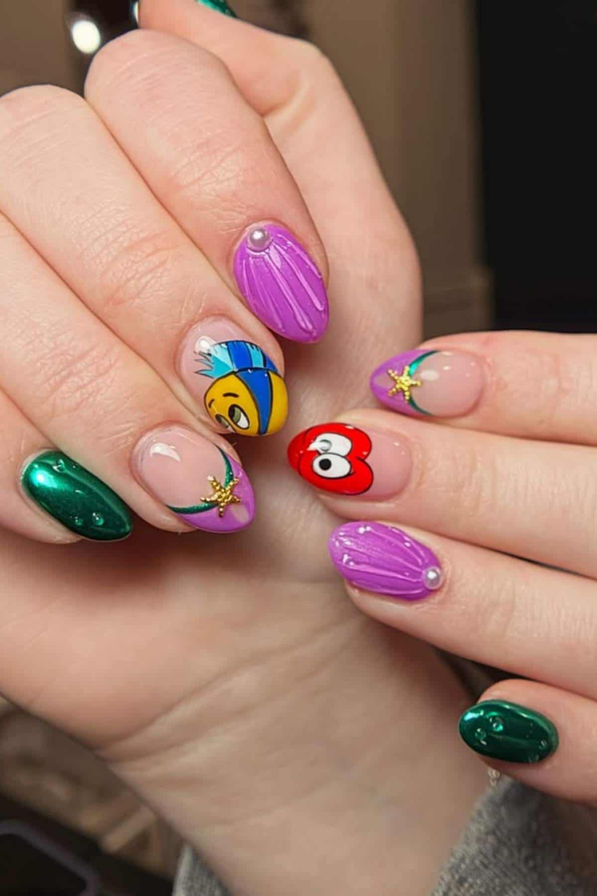 A hand with manicured nails featuring green, purple, and clear designs, some with pearls, flowers, and cartoon face accents.