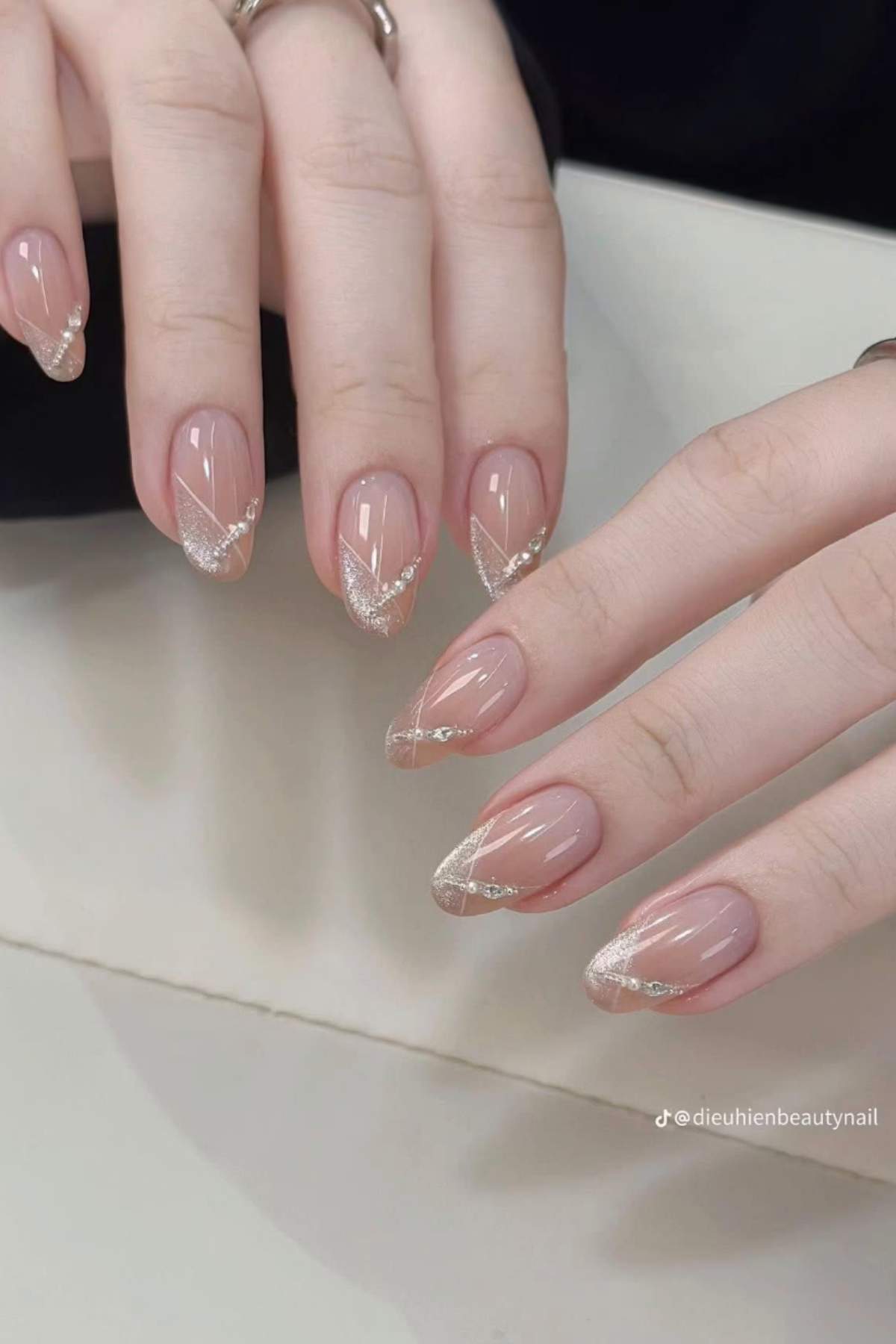 A close-up of hands with almond-shaped nails featuring a nude base and silver glitter diagonal tips.