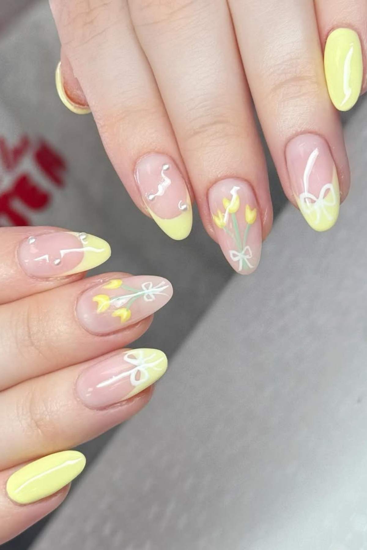Close-up of hands with almond-shaped nails featuring yellow French tips, white line art, and small yellow flower designs.