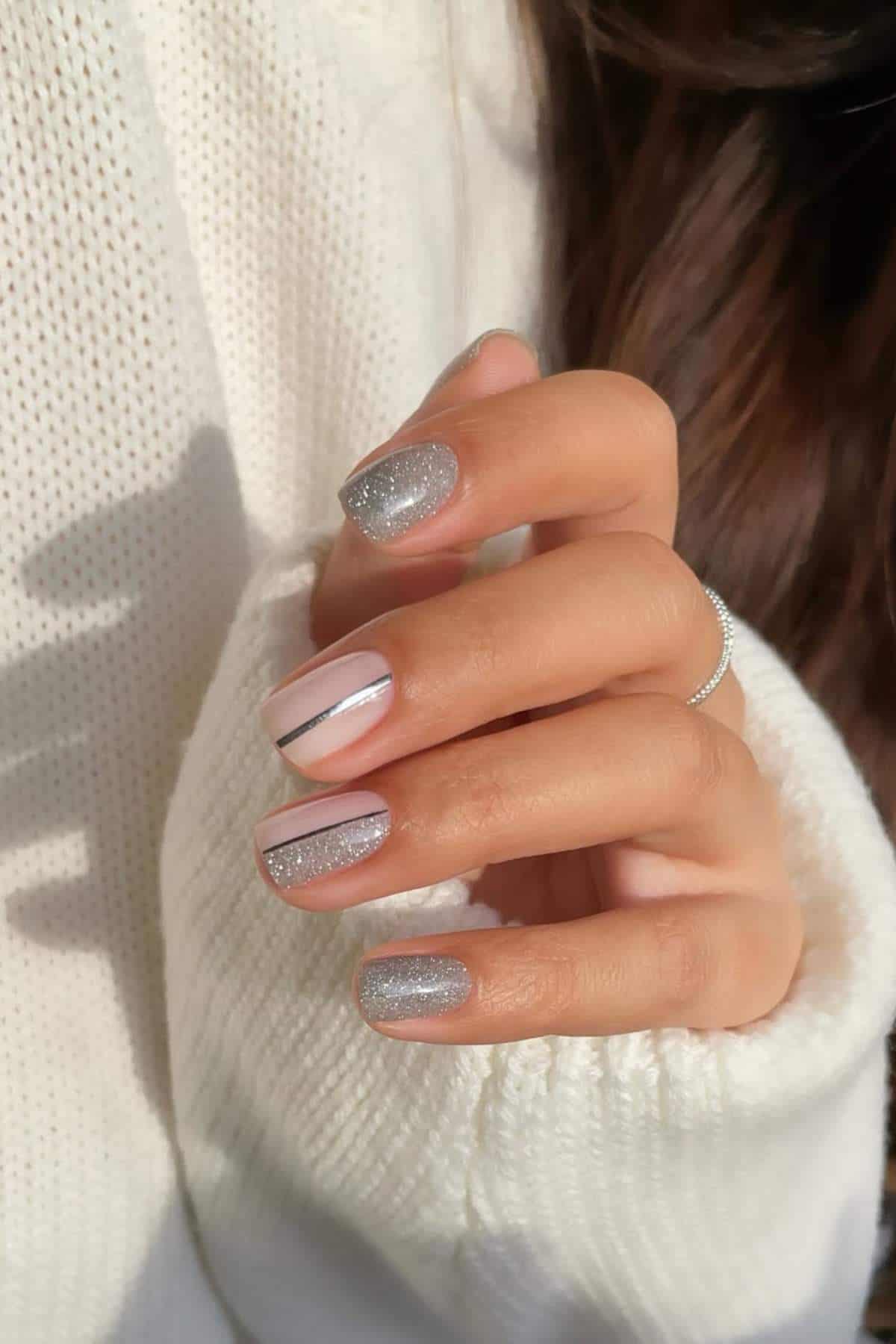A hand with manicured nails featuring silver glitter polish, light pink polish with a metallic stripe, and a thin silver ring on the index finger, against a white sweater background.