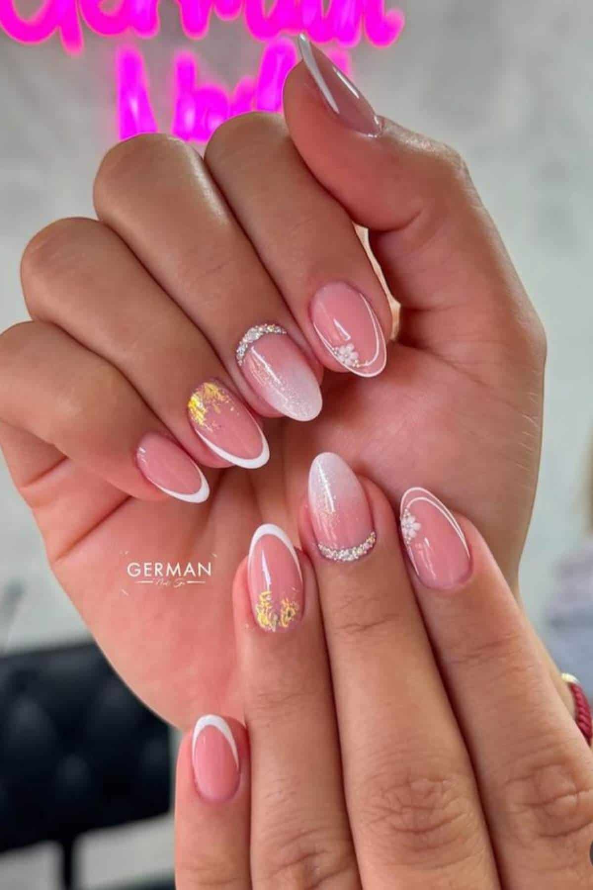 A close-up of hands with almond-shaped nails featuring a pink base, white French tips, gold foil accents, and delicate white floral nail art designs.