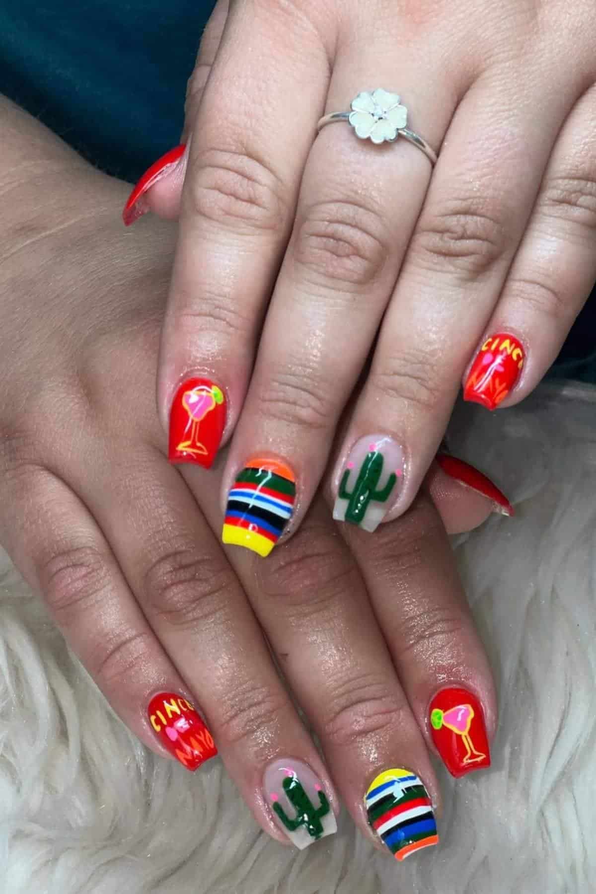 Hands with brightly colored nail art featuring cacti, margarita glasses, horizontal stripes, and the word "Cinco" on a couple of nails. A flower-shaped ring is on one finger.